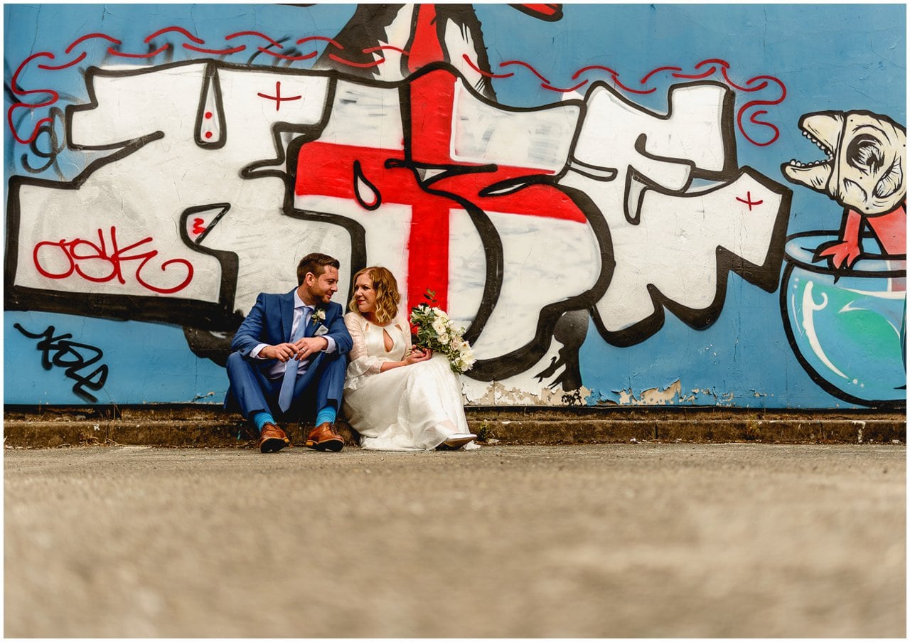 Bride and groom sitting infant of a big graffiti wall at The Custard Factory on their wedding day.