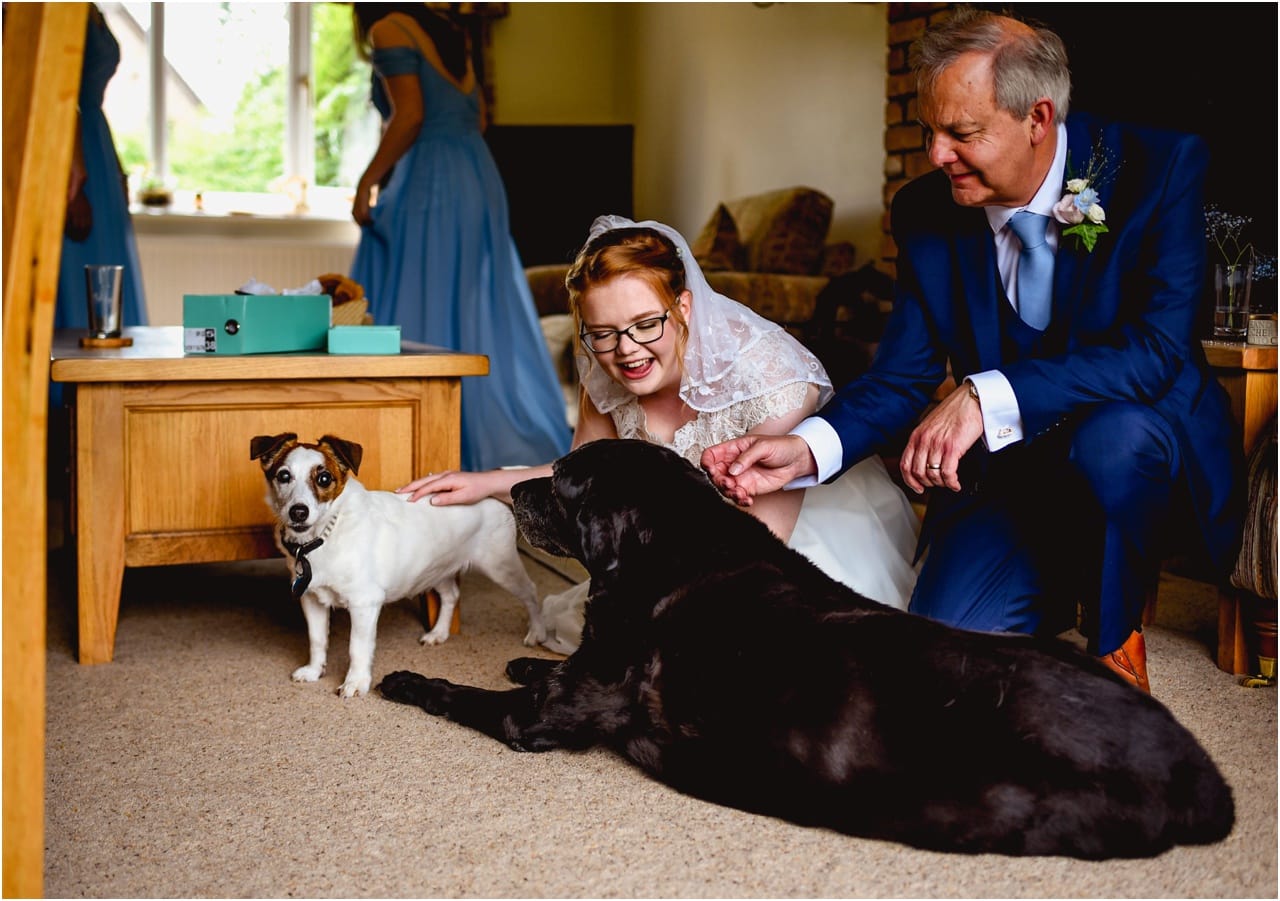 Bride and her Dad cuddle their pet dogs before leaving for the wedding, bride is in a white wedding dress and Dad is wearing a blue suit and pale blue tie.