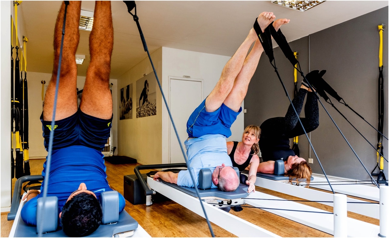 A reformer class using pilates machines, three men on their backs with their legs in the air.