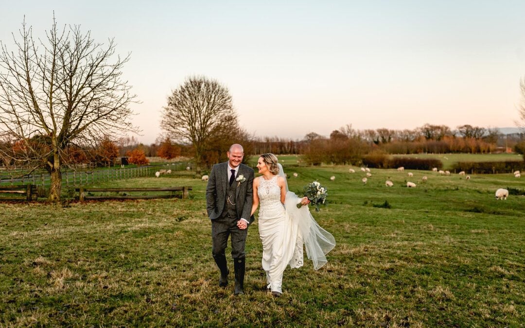 Bride and groom walk through a field at Mythe Barn Wedding Venue in their wellies surrounded by sheep.