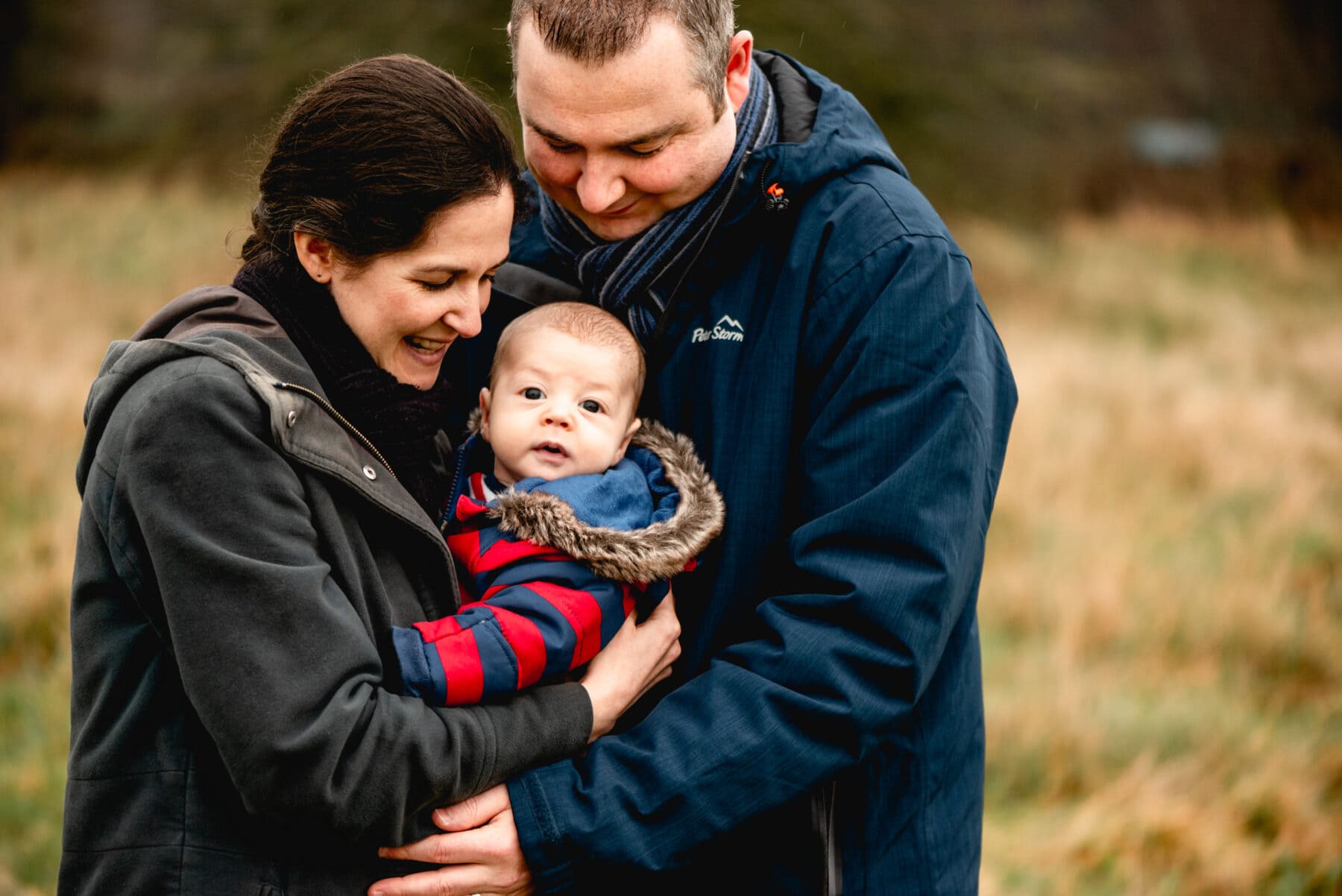 Family cuddle in the park during winter family shoot with newborn baby wrapped up warm in red and blue striped snowsuit.