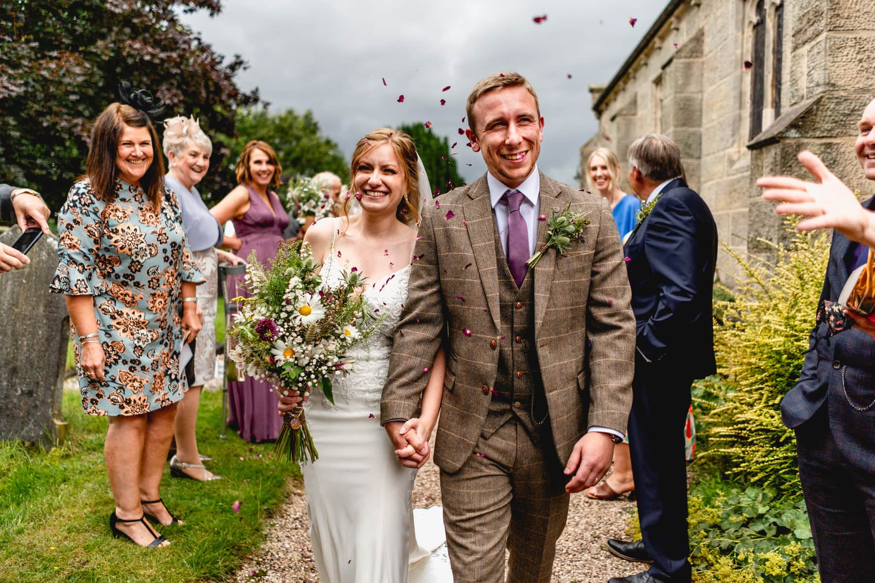 Bride and groom walk through a tunnel of confetti wearing a white dress and a beige tweed suit. Best Wedding Photography of 2021