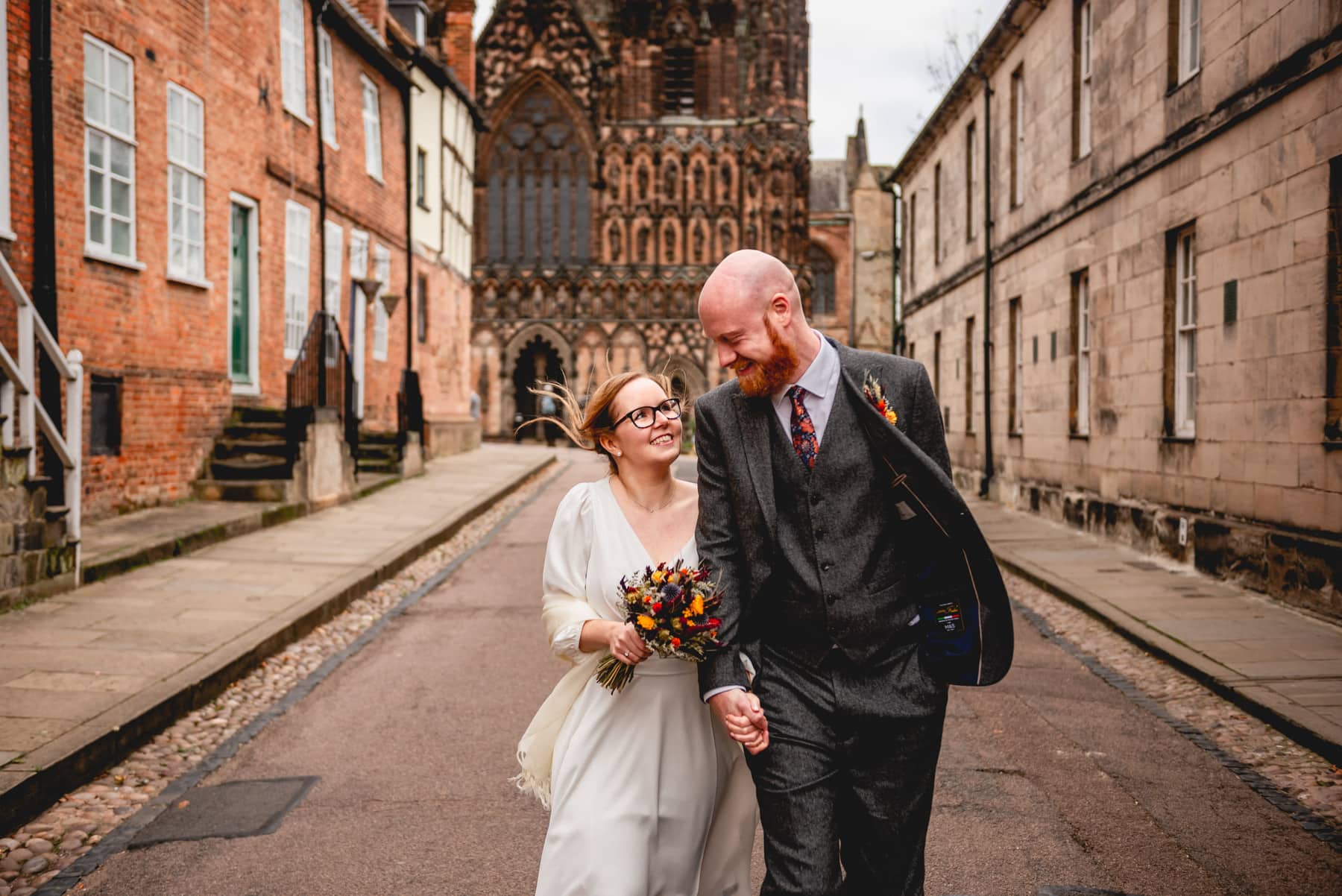 Bride and groom walk towards the camera looking at each other. There is a cathedral in the background and the pavements are cobbled. Bride wears glasses and white dress, groom wears grey tweed suit and patterned tie. Best Wedding Photography of 2021