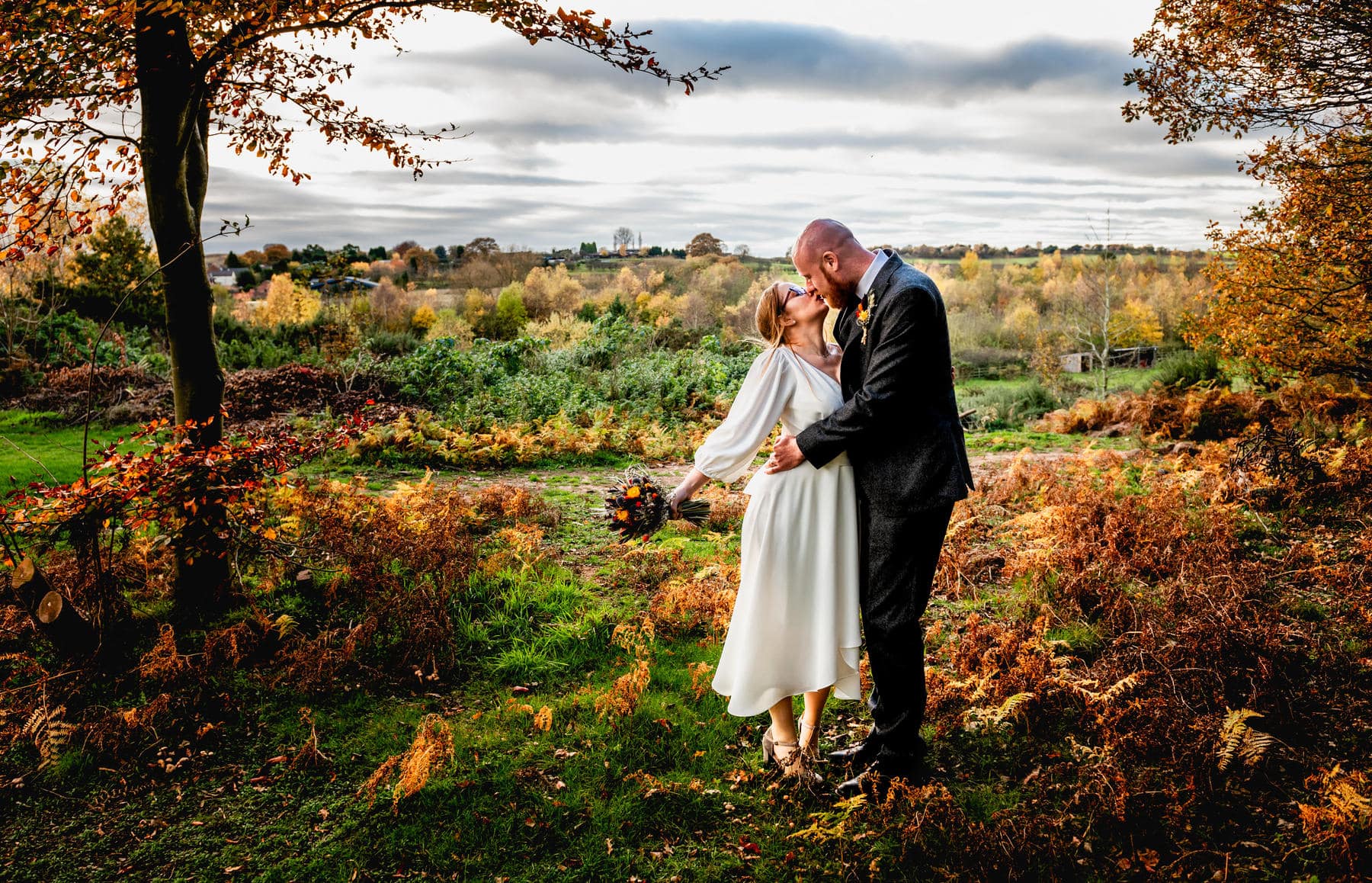 Bride reaches up to kiss her husband surrounded by autumn leaves wearing white dress and holding bouquet. Best Wedding Photography of 2021