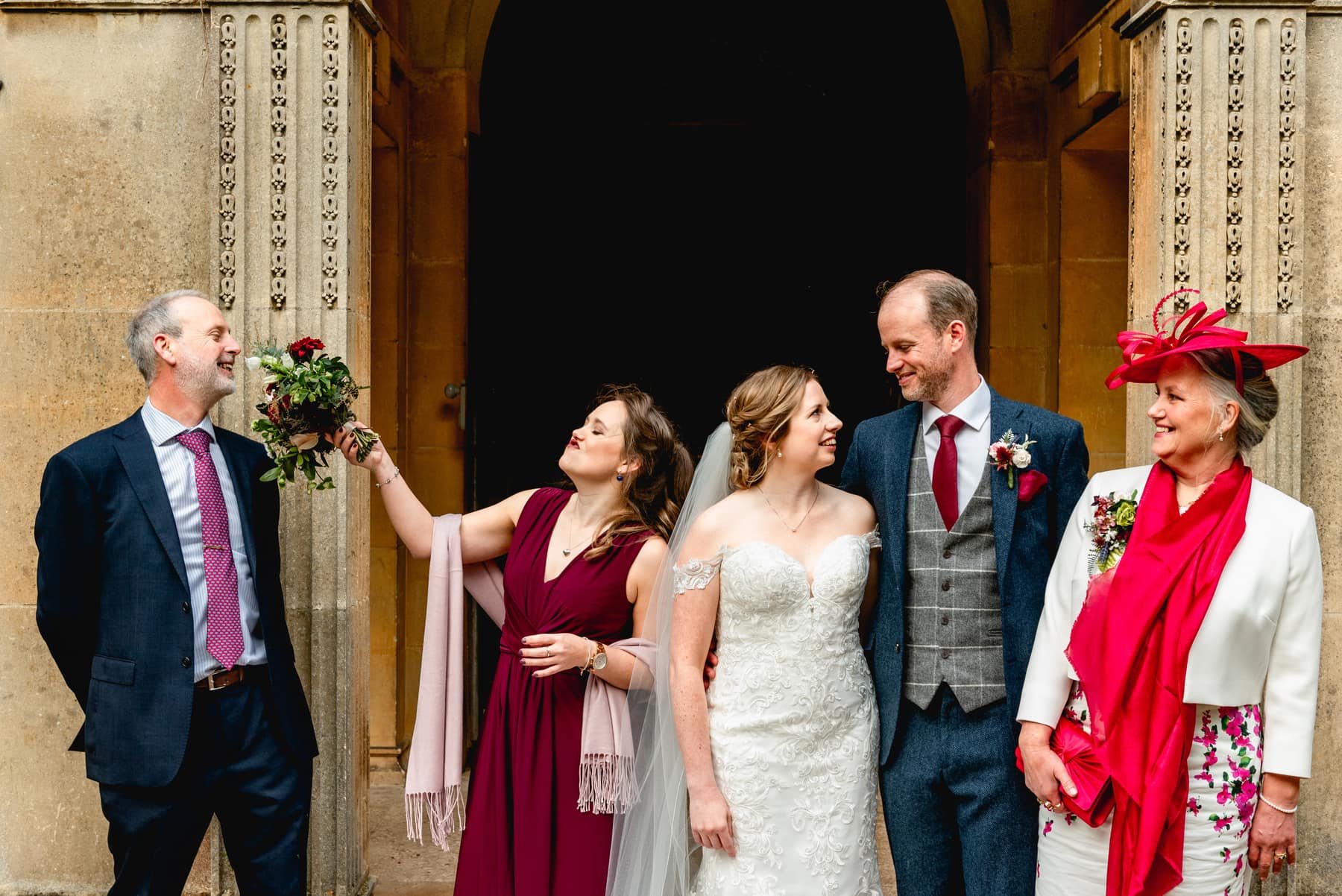 Family portrait at a wedding. Bridesmaid taps father of the groom on the nose with a bouquet while mother watches on laughing and bride and groom smile at each other. Best Wedding Photography of 2021