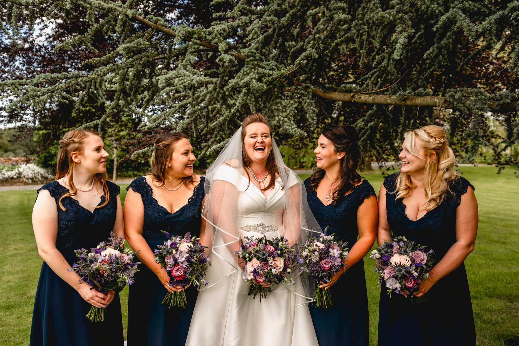 Bridesmaid squad cuddle together for a photo, all laughing and all holding bouquets, bride wears white wedding dress and bridesmaids wear long length navy blue lace dresses. Best Wedding Photography of 2021