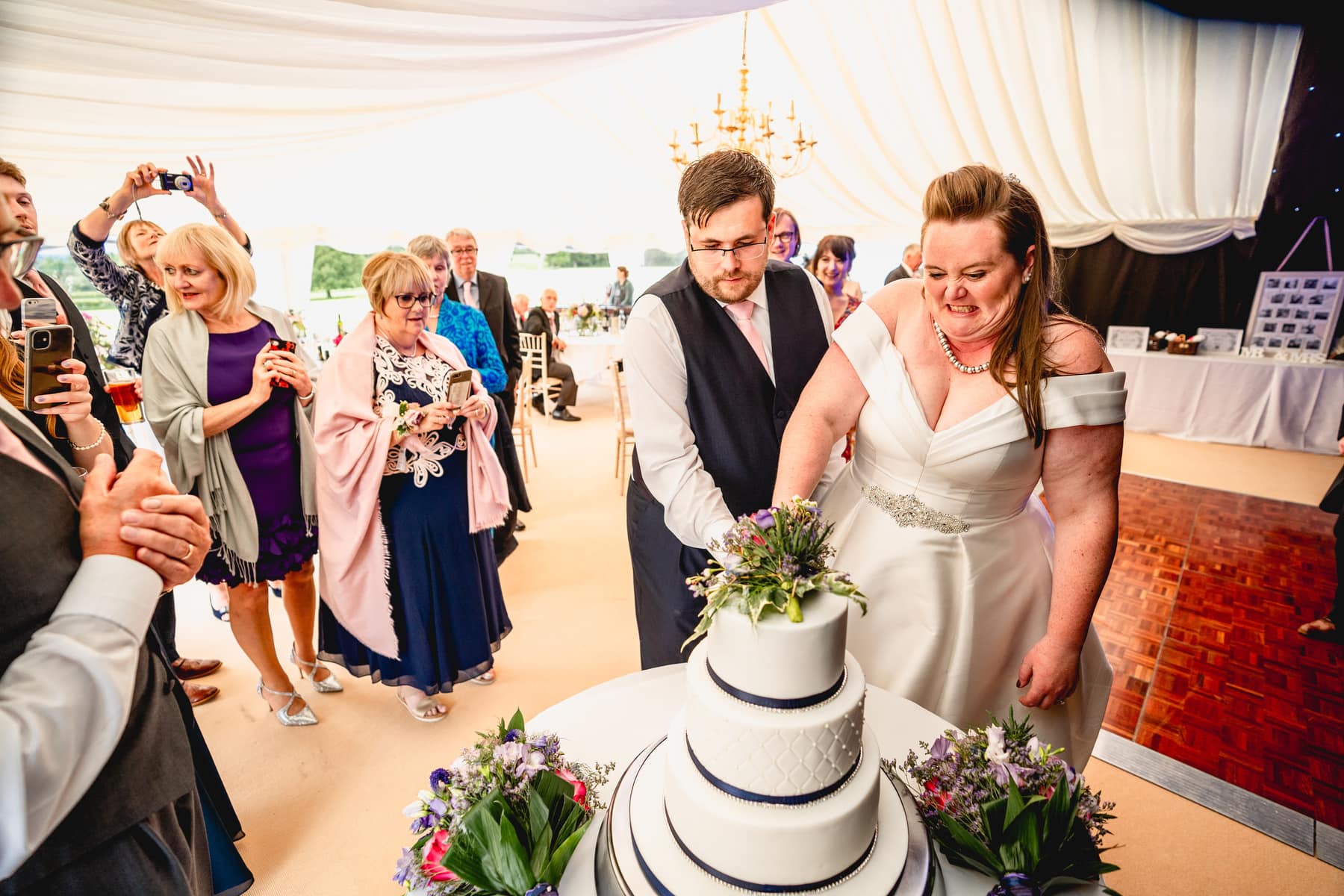 Bride and from grimace as they cut 4 tier wedding cake in a marquee