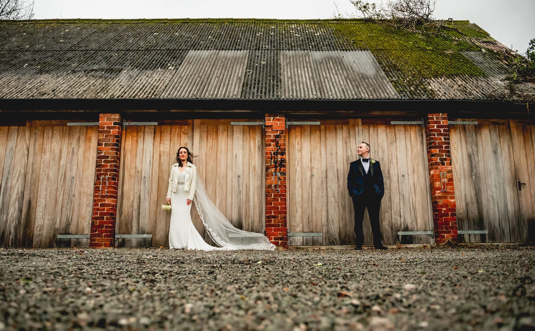 Bride and groom stand separately against wooden garage doors on a gravel path, brides veil blows in the wind and she hold a white rose bouquet. Best Wedding Photography of 2021