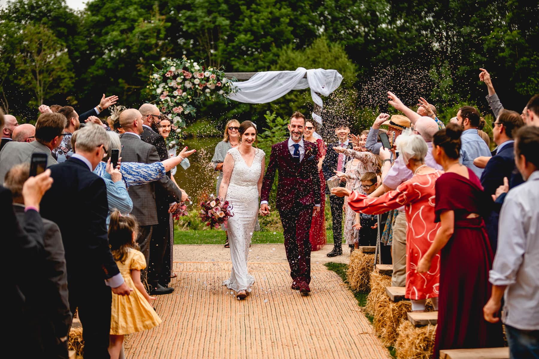 Couple exit their humanist celebration showered with confetti by a lake with a flower arch behind them. Best Wedding Photography of 2021