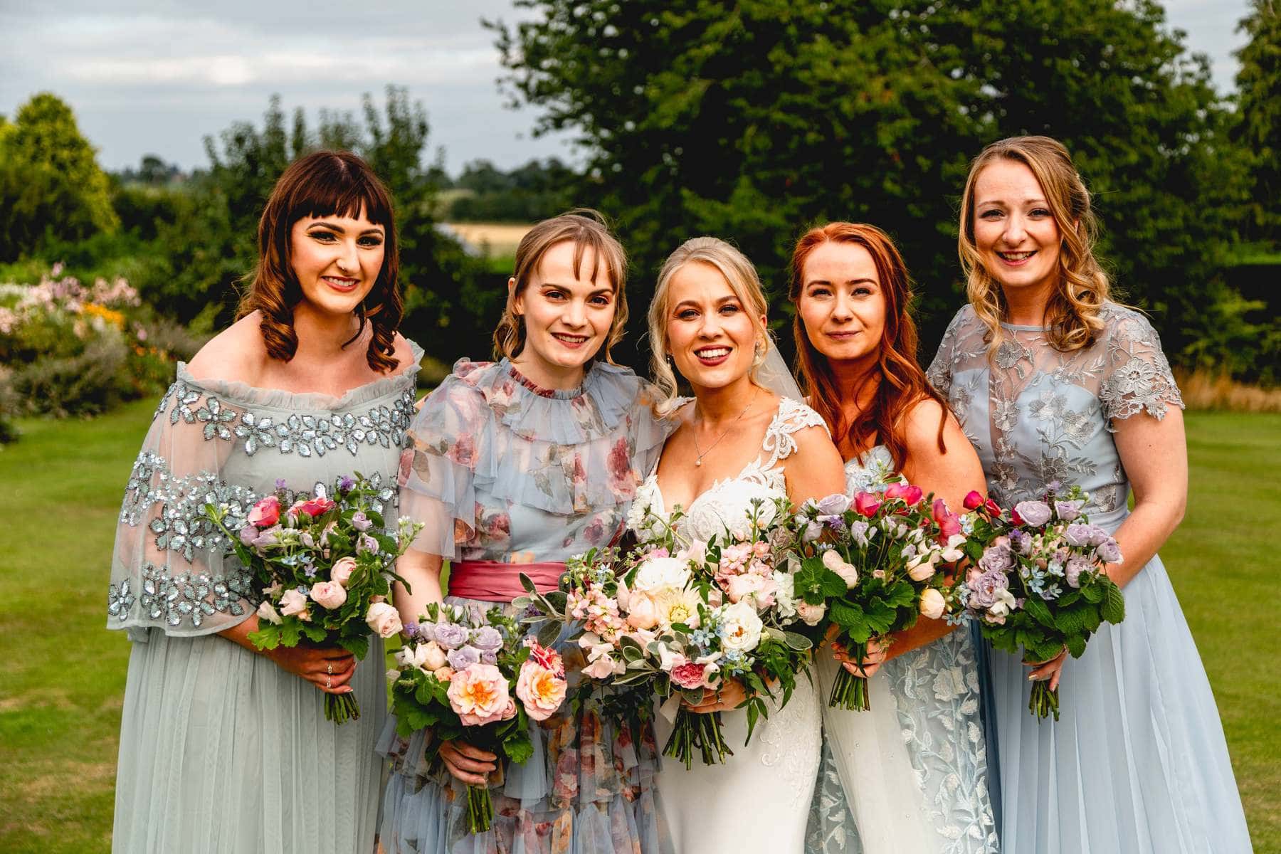 Bridesmaids all wear different tones of blue floral dress. Five girls all stand together with flower bouquets.