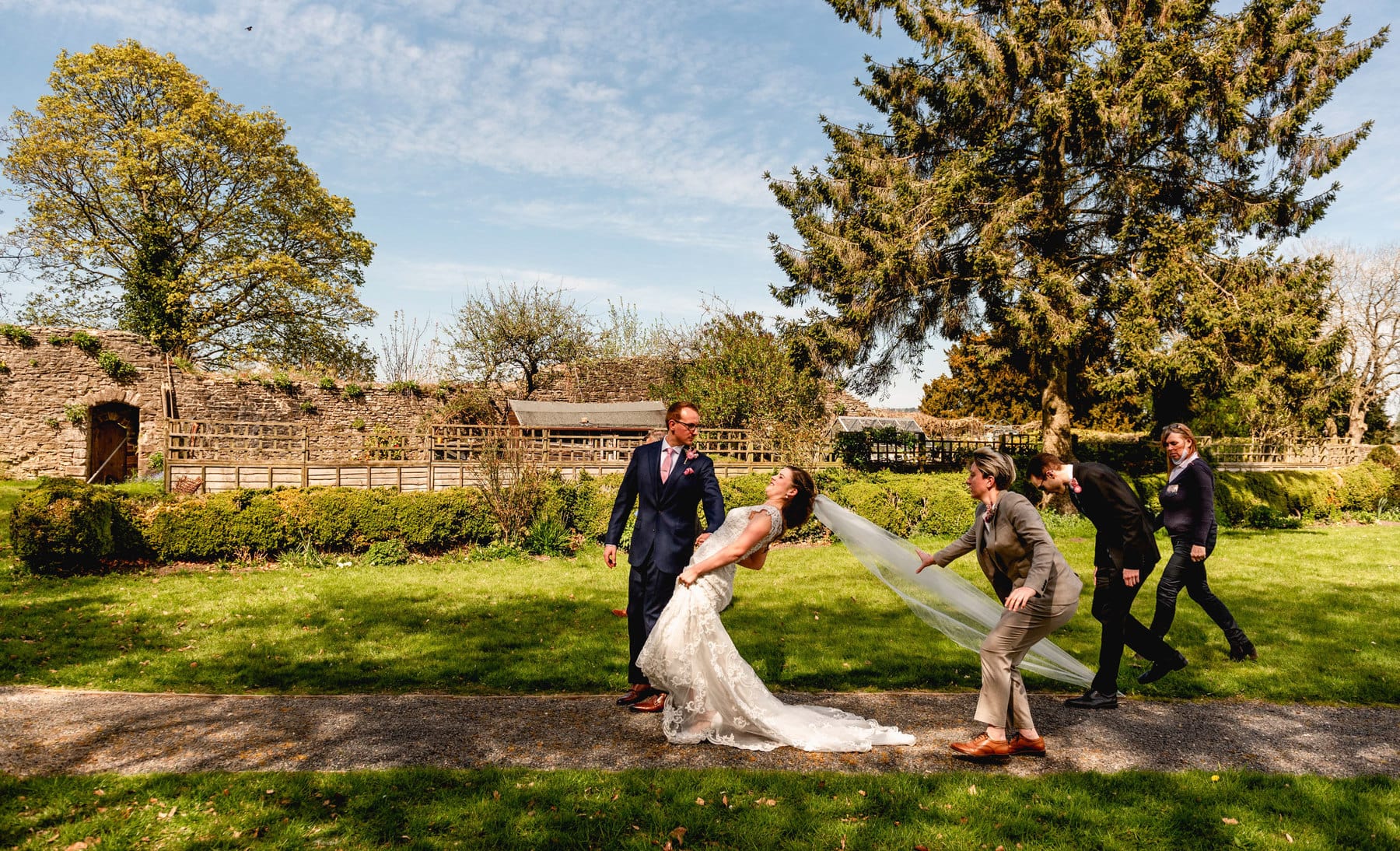 Award winning photo of bride pulled back as best man stands on her veil and bridal party all rush to help