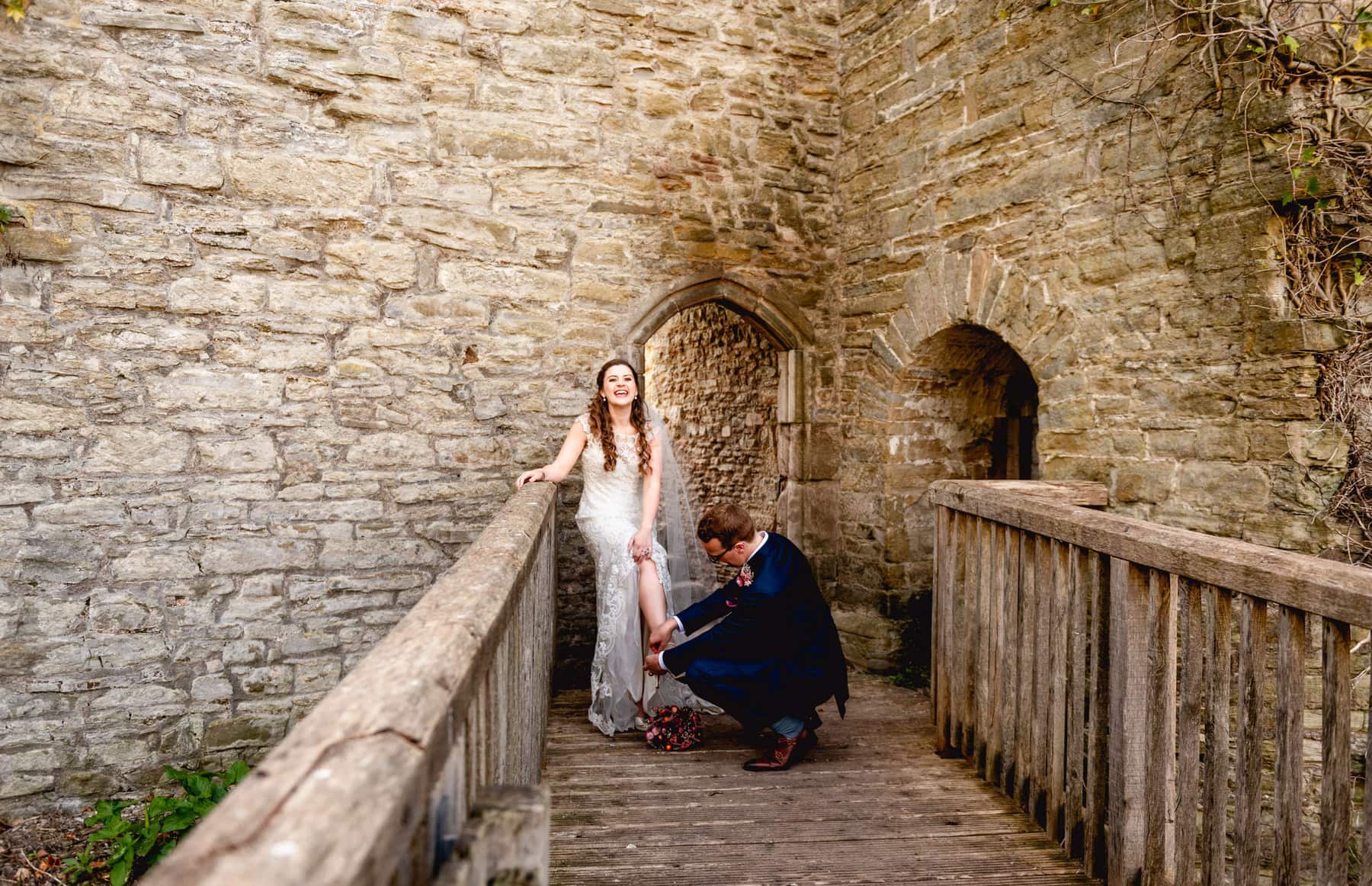 Bride laughs as groom crouches down trying to put her shoe back on on a wooden bridge at a castle