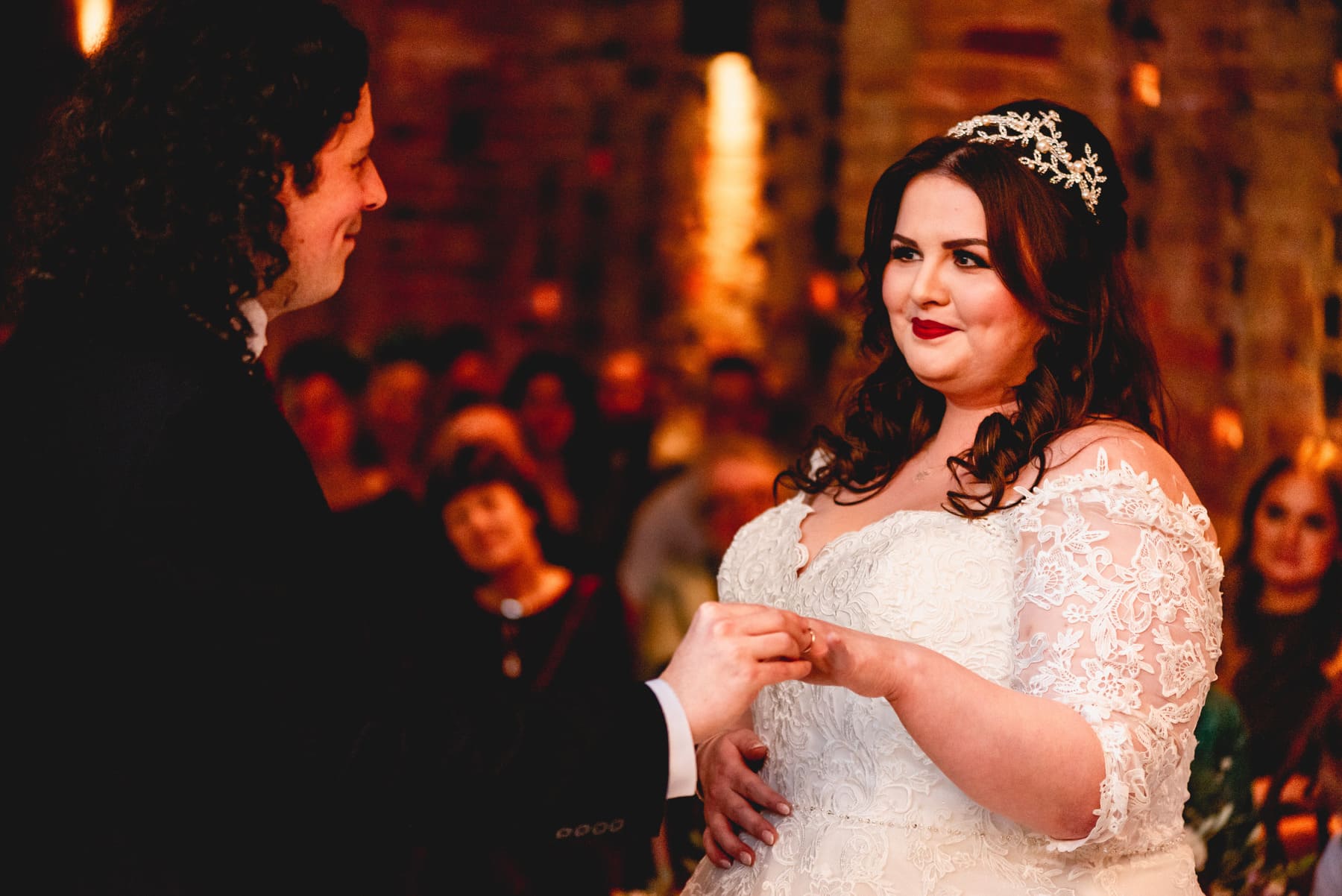 Bride smiles as new husband places ring on her finger. Red lipstick and hair crown.