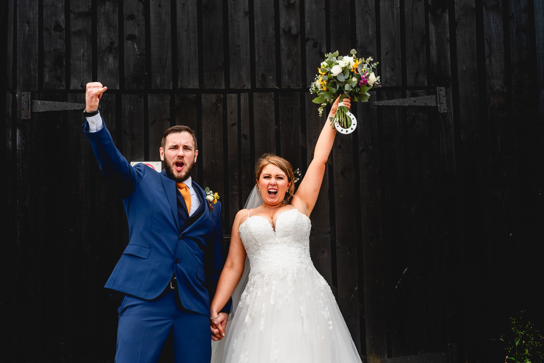 Couple punch the air after getting hitched at Curradine Barns standing a black barn door. Bride holds bouquet and horseshoe in the air. Best Wedding Photography of 2021. Best Wedding Photography of 2021