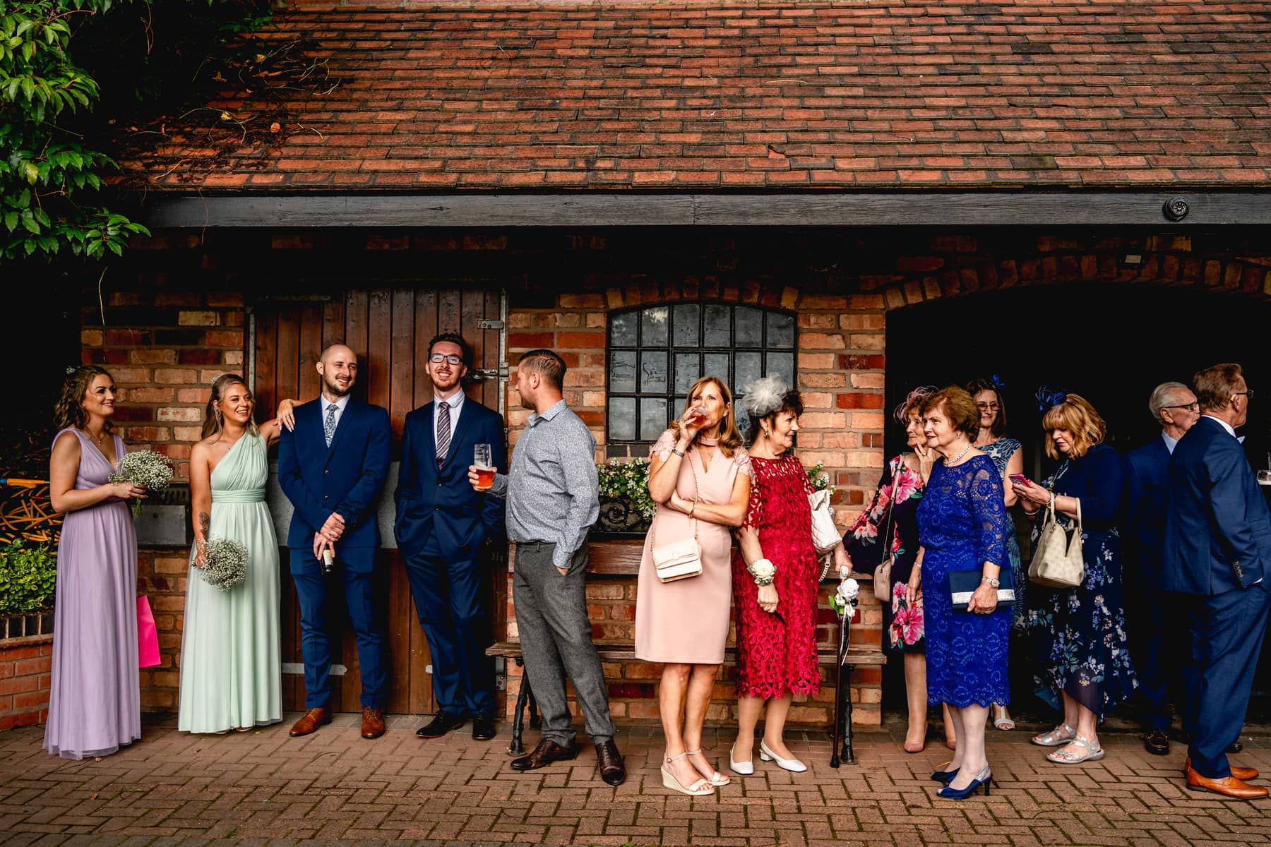 Wedding guests stand around having a drink against a brick barn chatting to each other. candid photography. Best Wedding Photography of 2021