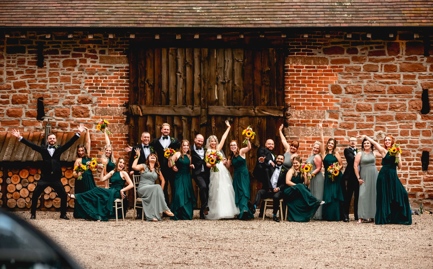 Large bridal party all strike a funny pose together. Women wear dark green dresses and sage green long length gowns with men wearing black dinner jackets and dickie bows. Women all hold sunflower wedding bouquets