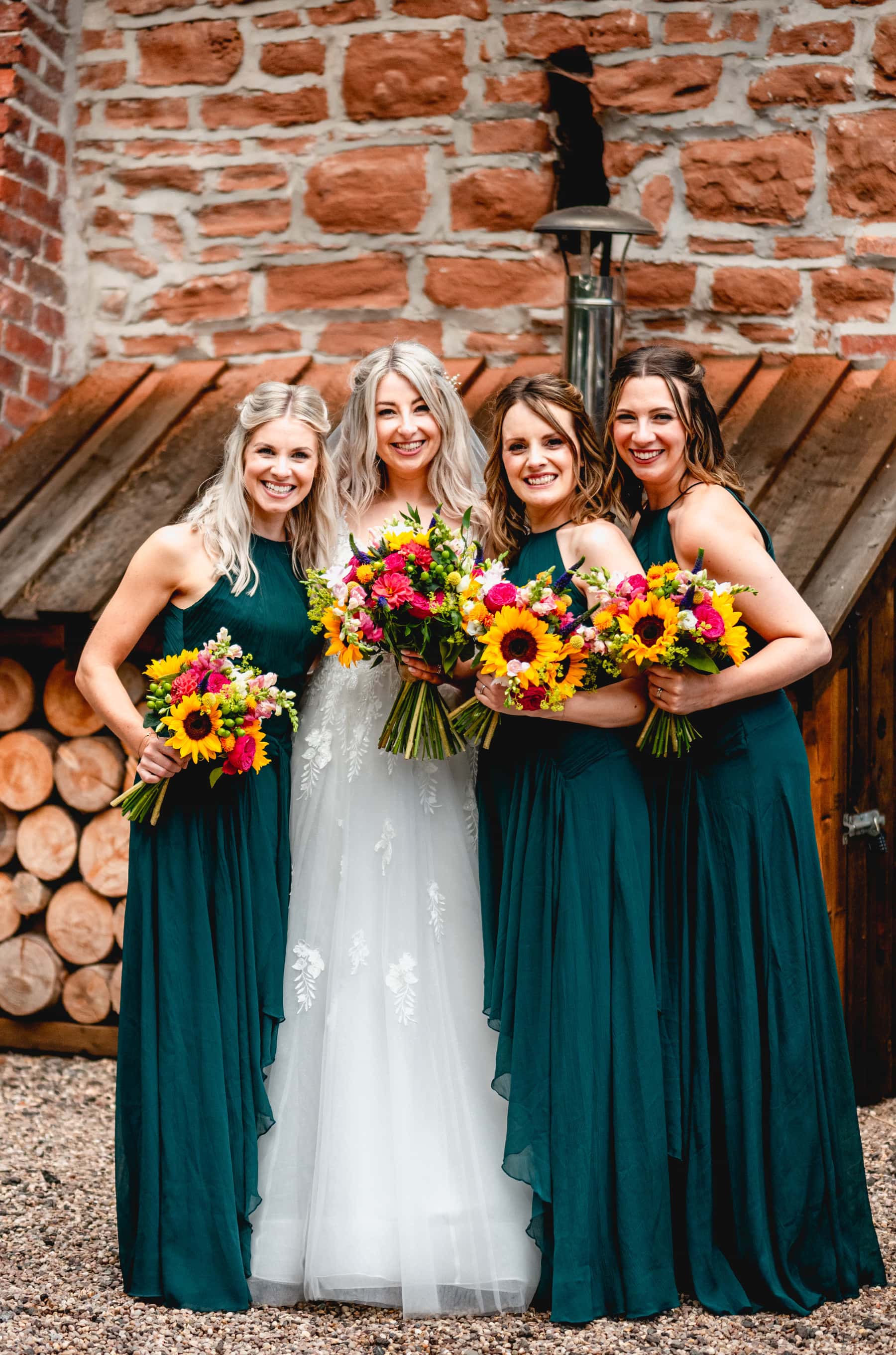 Bride and bridesmaids stand closely together infant of logs wearing jade green long length dresses and holding bouquets with sunflowers. Best Wedding Photography of 2021