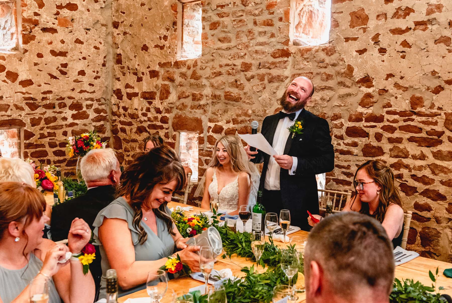 Groom wearing dinner jacket and yellow flower buttonhole stands to make his speech holding a microphone inside a stone barn