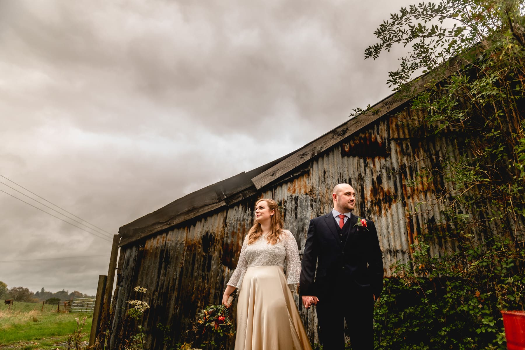 Bride in cream long sleeved wedding dress holds bouquet and groom holds her hand standing against a rusted corrugated iron backdrop. Best Wedding Photography of 2021