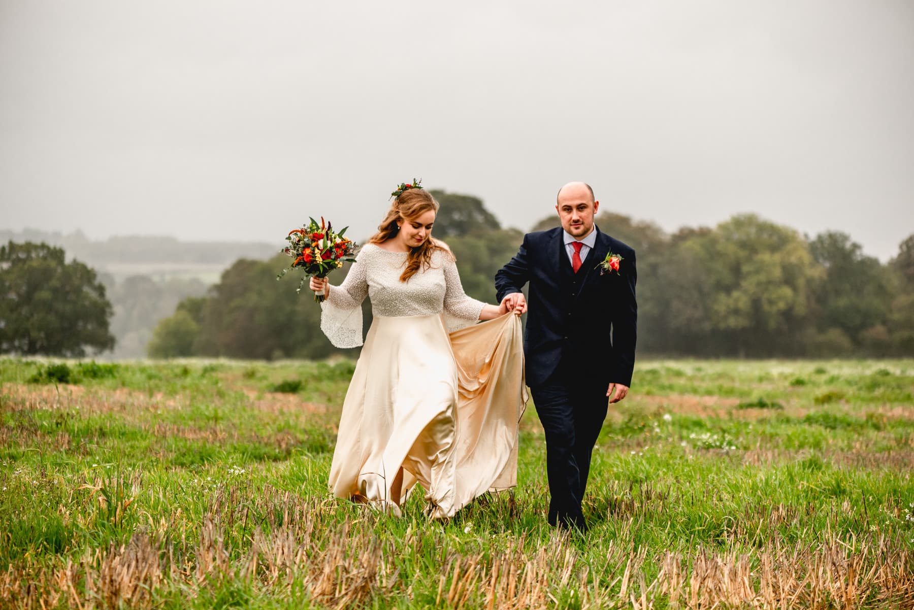 Bride and groom walk through field after their wedding, groom holds brides skirt to keep it from the floor, bride holds bouquet
