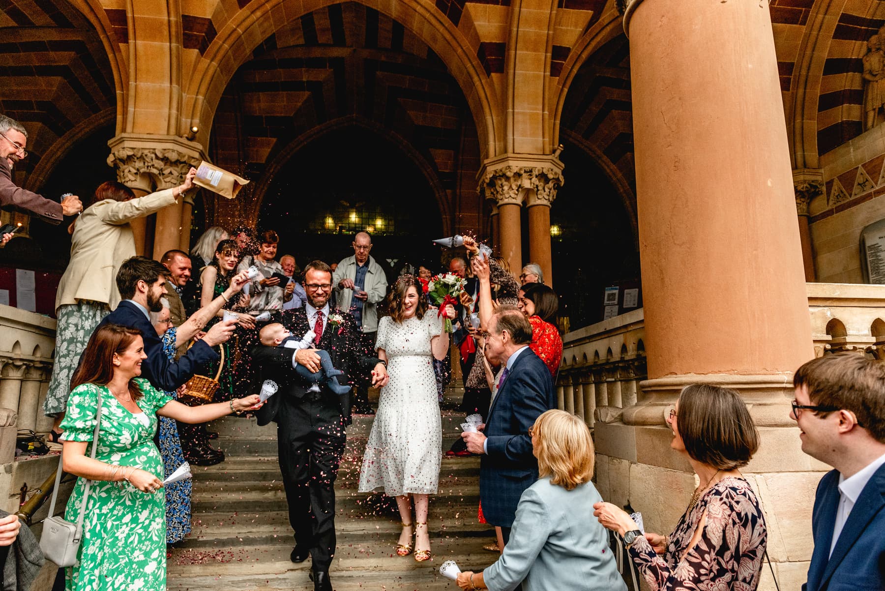 Newly married couple walk down stairs of registry office in Northampton showered in confetti while carrying a baby. Bride wears white tea dress with gold polkadots. Best Wedding Photography of 2021