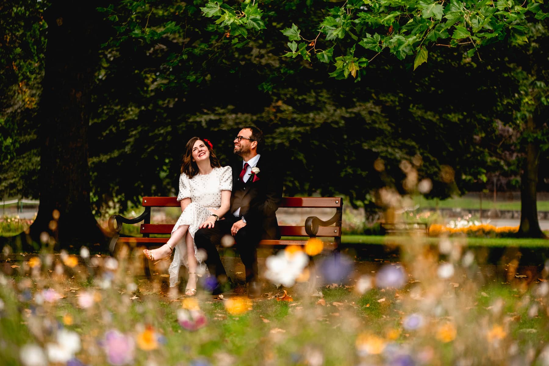 Couple sit on a bench in a park surrounded by wildflowers