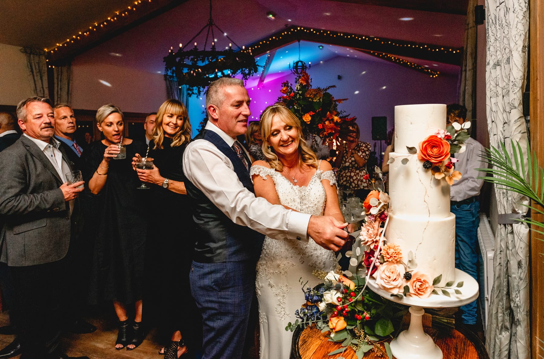 Couple cut the wedding cake while guests look on. Cake is three tier with orange and peach floral accents. Best Wedding Photography of 2021