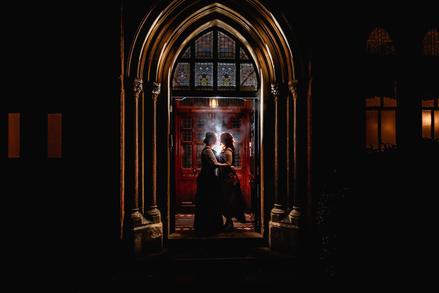 Two brides face each other in the doorway of a stately home at night-time for an evening portrait backlit by a starburst of flash and some fog. Doorway architecture and stained glass windows. Best Wedding Photography of 2021