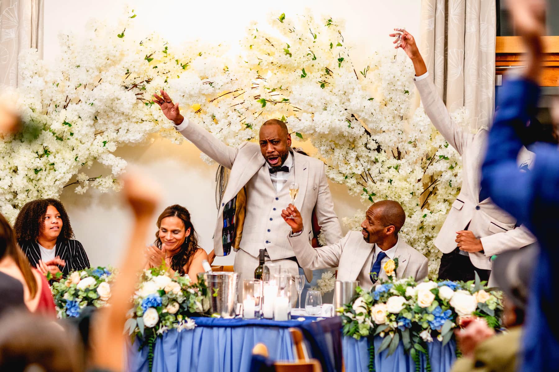 Groom makes a speech at his wedding top table wearing creme suit and black tie with waistcoat. Guests salute with glasses of champagne. Best Wedding Photography of 2021