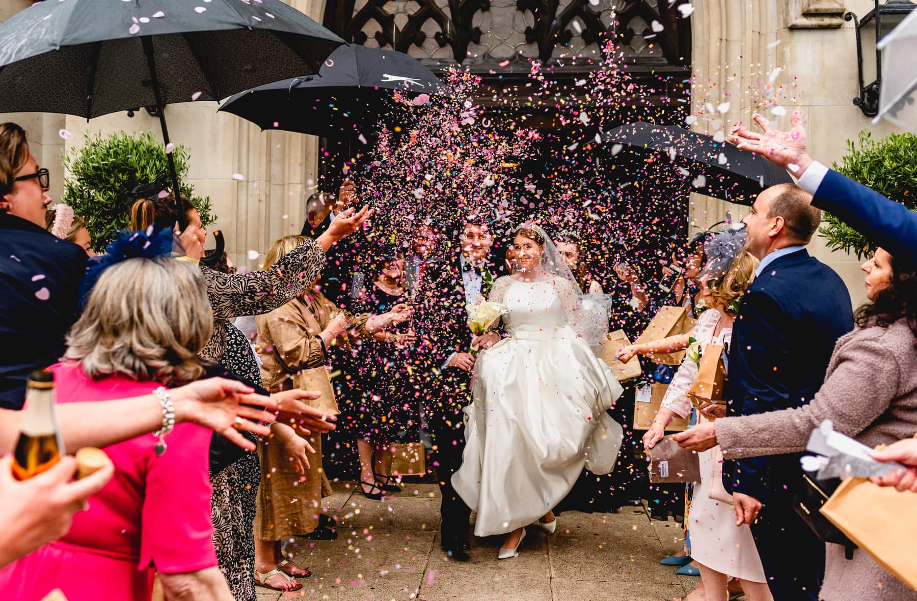 Newly married couple walk through shower of pink confetti during a rainy wedding in London at Southwark Cathedral doors. Best Wedding Photography of 2021