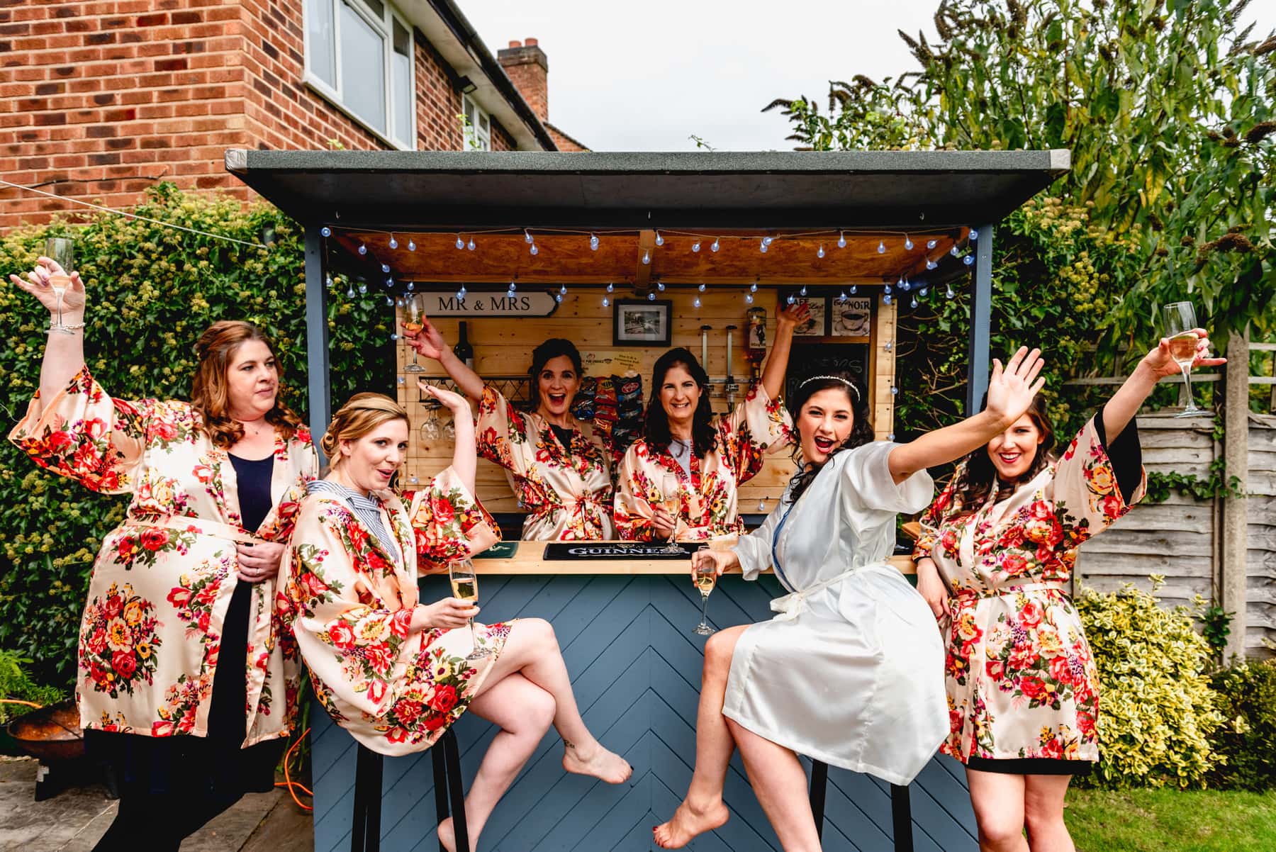 Bridesmaids all sit in garden during bridal preparation wearing floral dressing gowns with glasses of champagne at outdoor bar. Best Wedding Photography of 2021