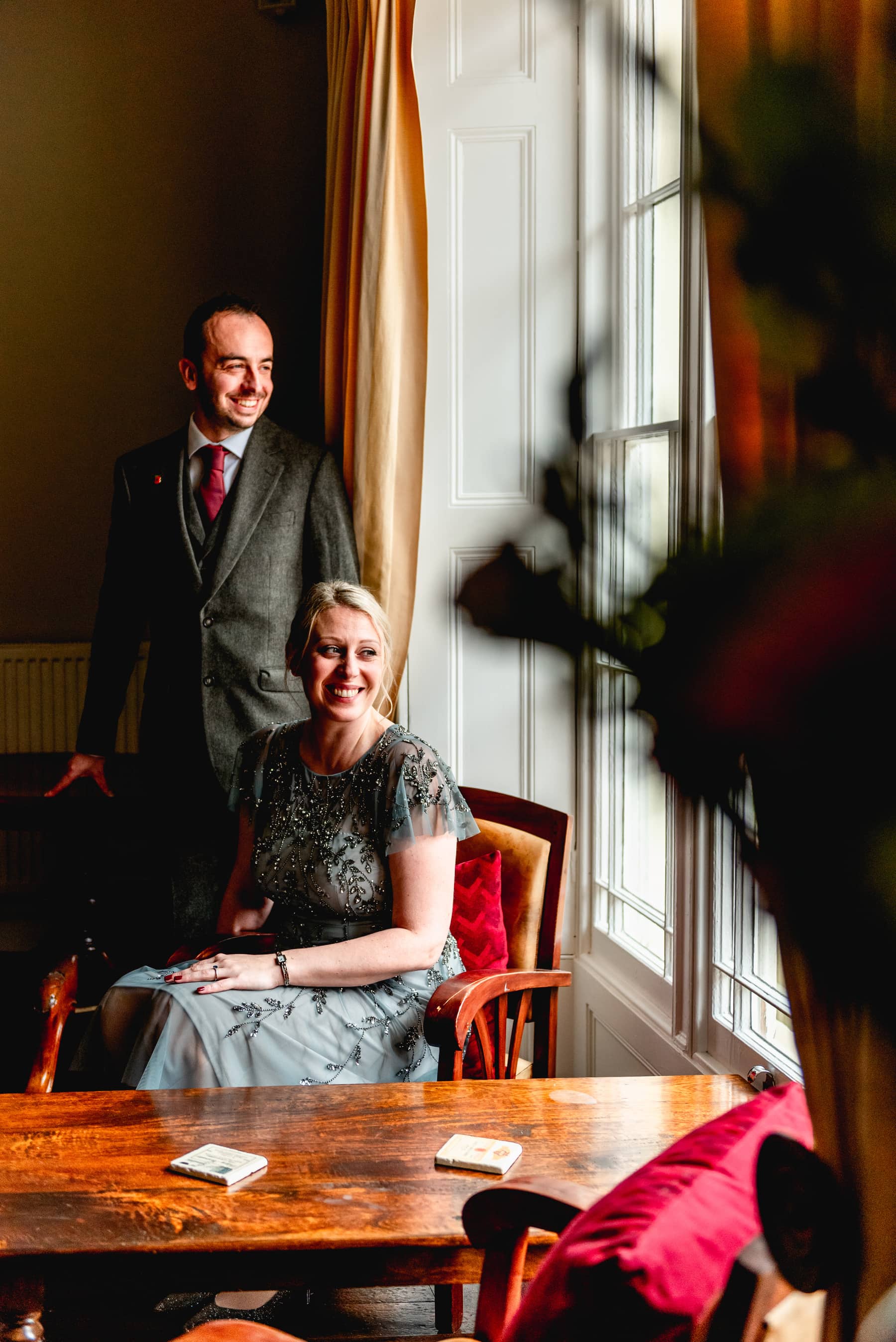 Bride in grey Monsoon dress sits in window of St Johns House with n ew husband wearing brown tweed suit and red tie