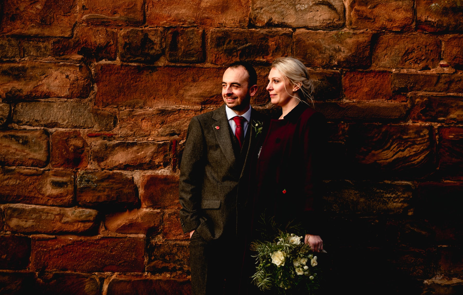 Bride in grey Monsoon dress and red jacket stands with new husband wearing brown tweed suit and red tie outside bridesmaids-a-brac shop. Best Wedding Photography of 2021