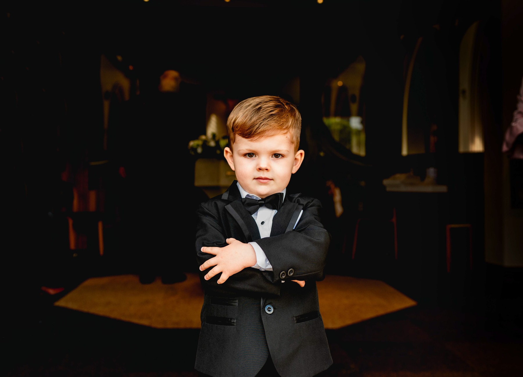 Page boy stands in church doorway wearing dinner jacket and bowtie with arms folded like a bounce