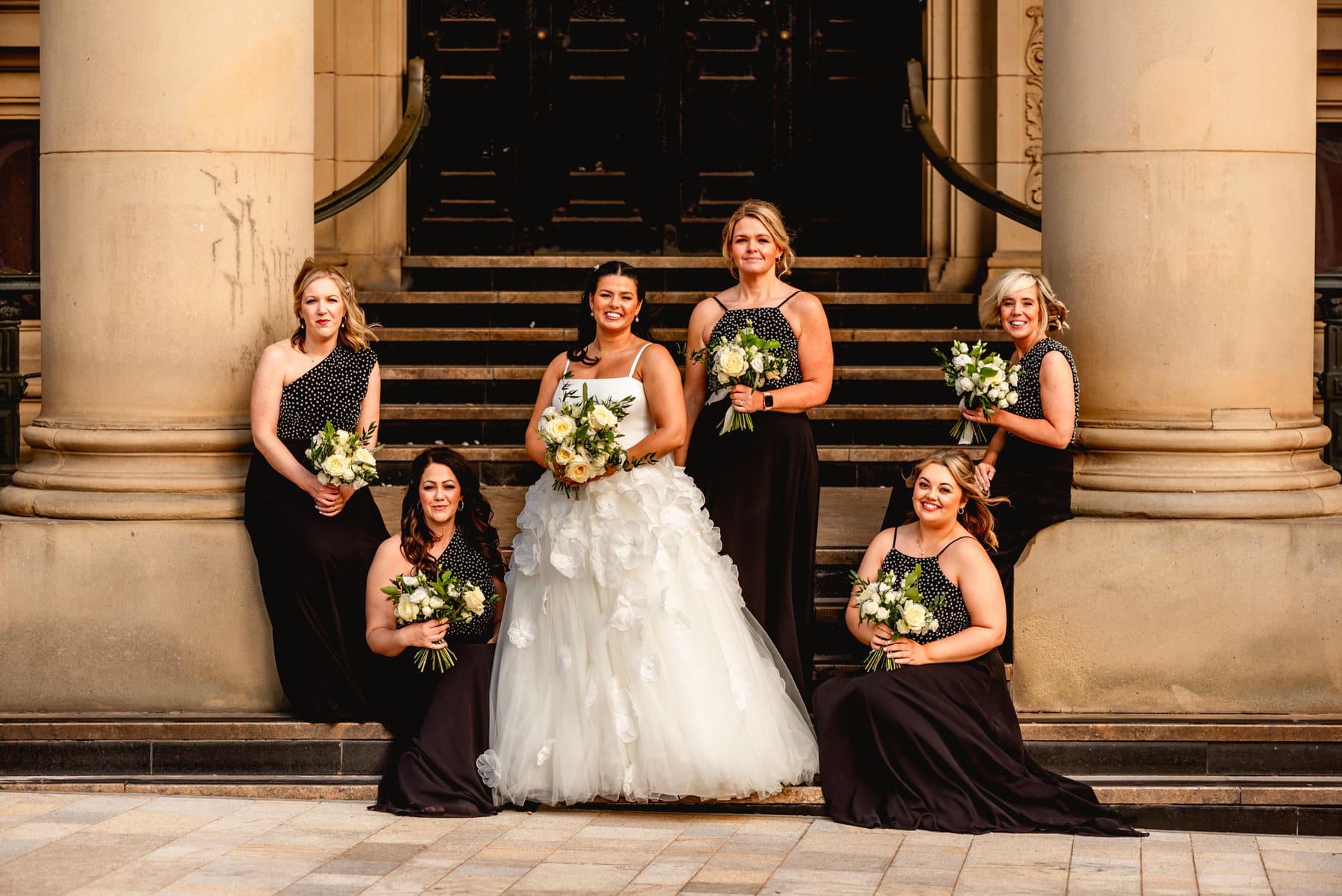 Bride and her bridesmaids on the steps of an art gallery on a wedding day, all hold yellow rose bouquets, bridesmaids are dressed in black dresses with pearl detail