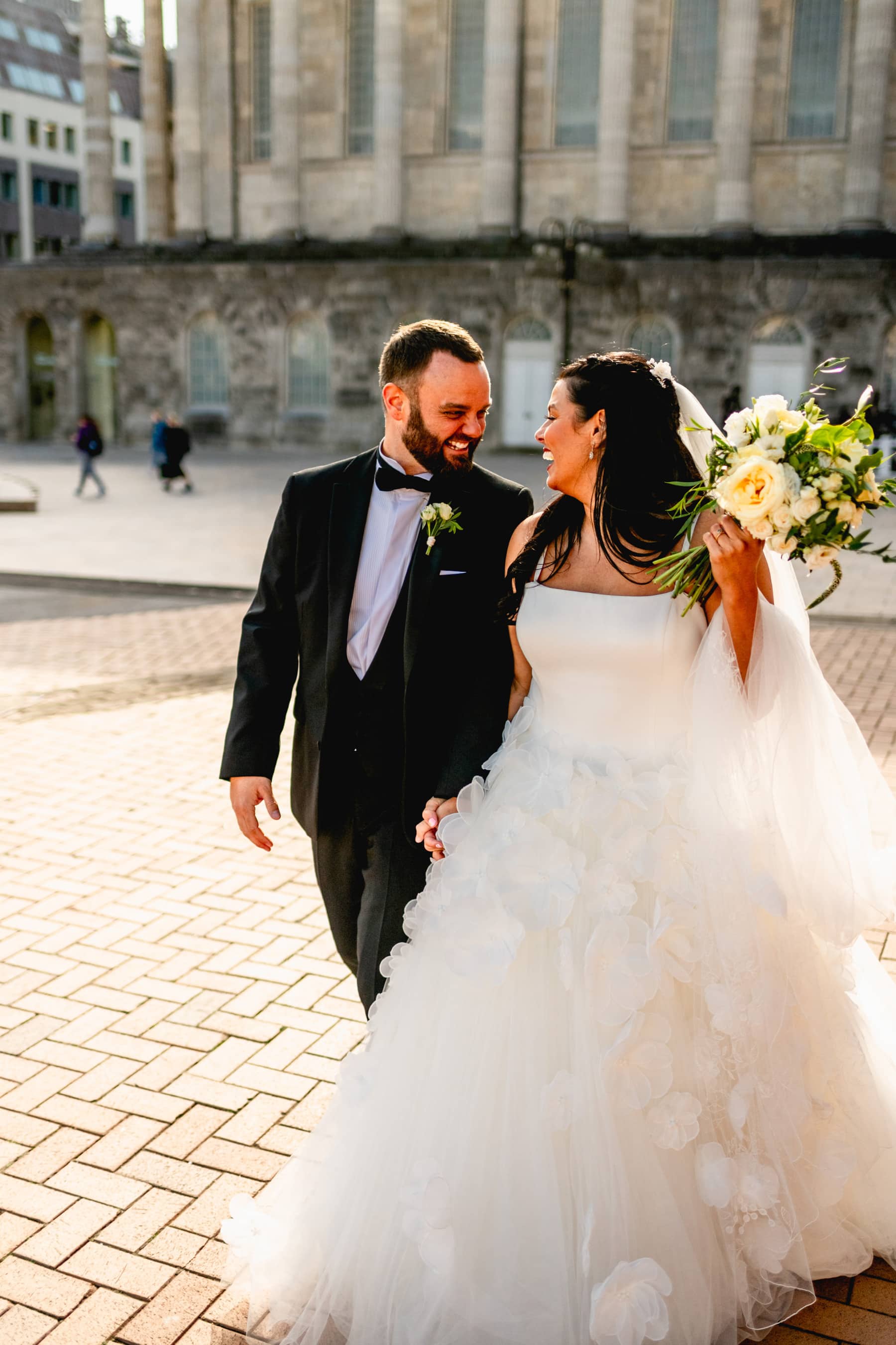 Bride and groom walk through the streets of Birmingham smiling at each other and holding a yellow rose bouquet. Groom wears black dinner jacket and bowtie