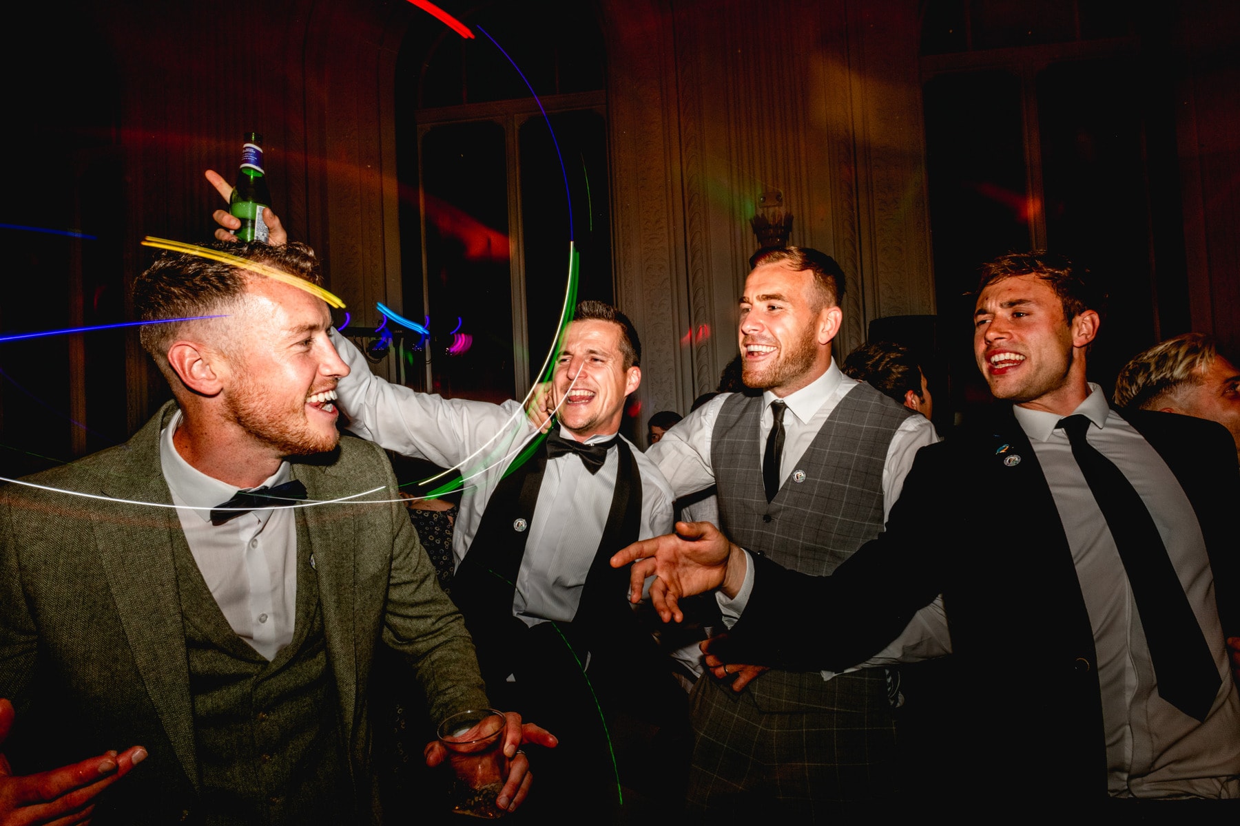 Wedding guests party with bottles of beer on the dance floor wearing suits and wedding favour pins.