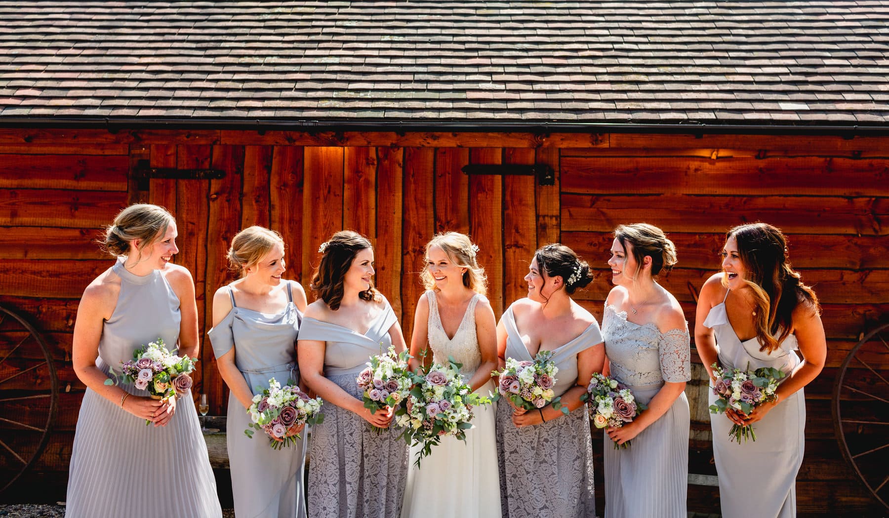 Bride and her six bridesmaids, all wearing grey dresses in different styles looking at each other and laughing, holding bouquets.