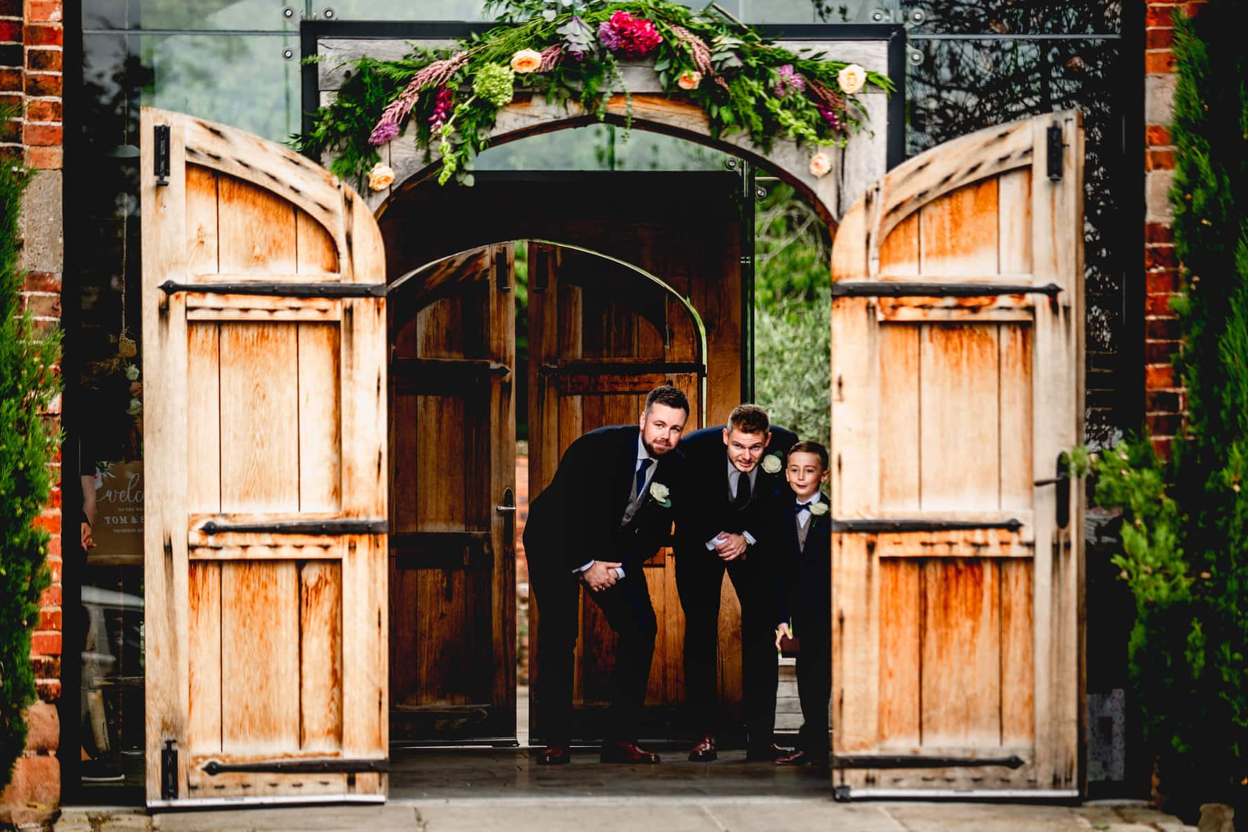 Groomsmen and page boy stand in doorway of barn wedding venue trying to see the brides arrival.