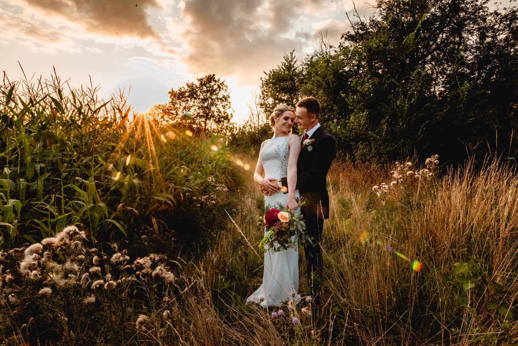 Couple stand in a field wearing wedding dress and suit as sun sets behind them at golden hour. Best Wedding Photography of 2021