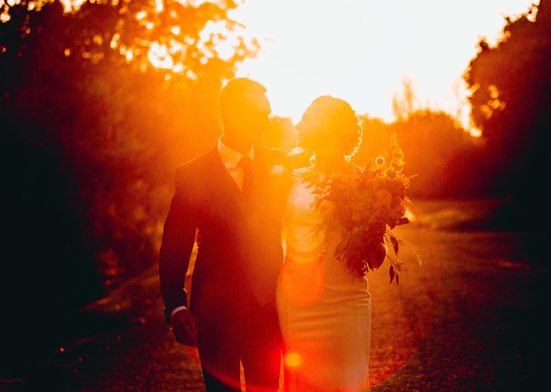 Bride and groom in hazy golden sunlight walk down a country path holding a bouquet on their wedding day.
