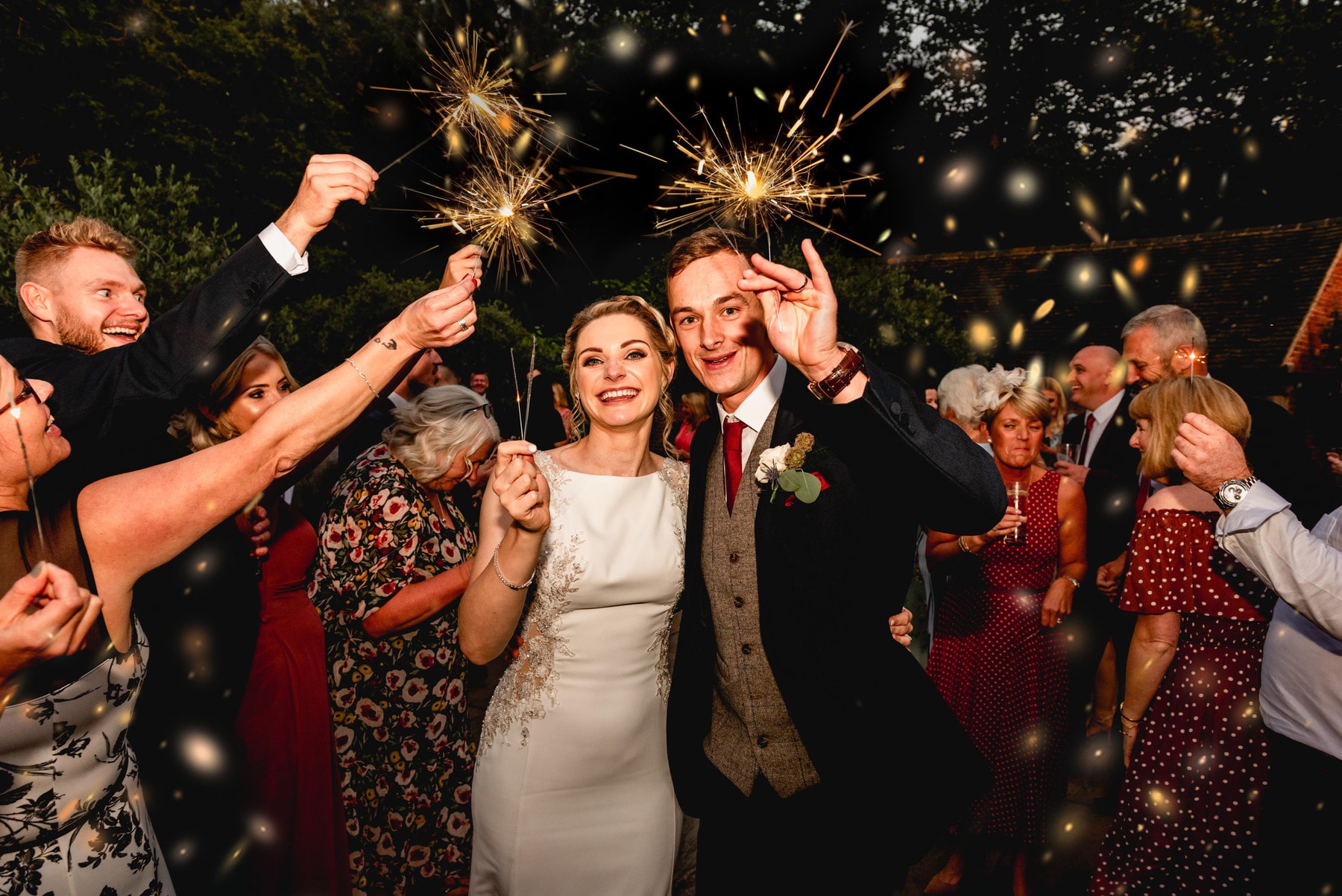 Bride and groom hold sparklers in the air as guests cheer them on with a sparkler tunnel.