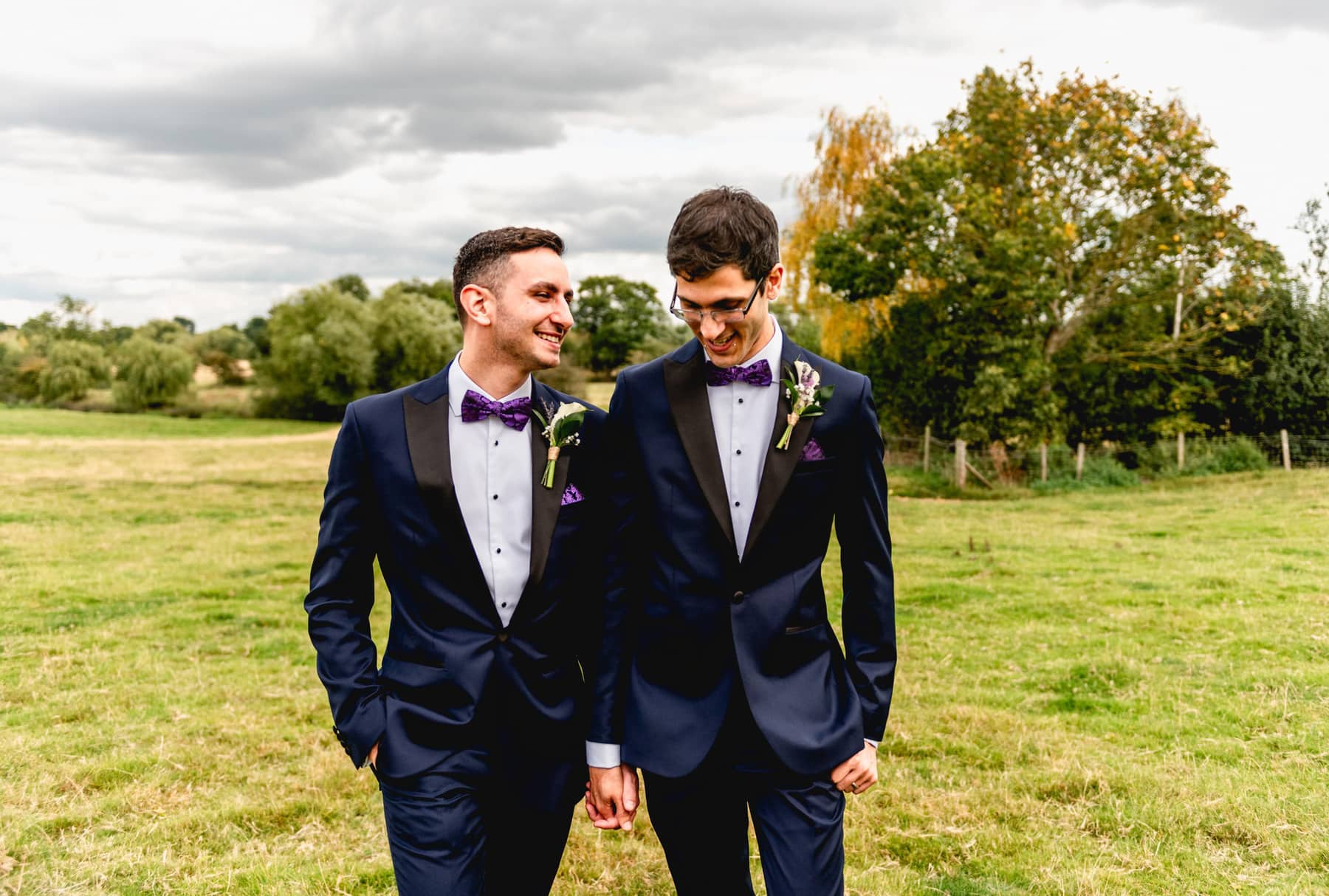 Gay wedding with two men in blue dinner jackets wearing purple bowties walking through a field. Best Wedding Photography of 2021
