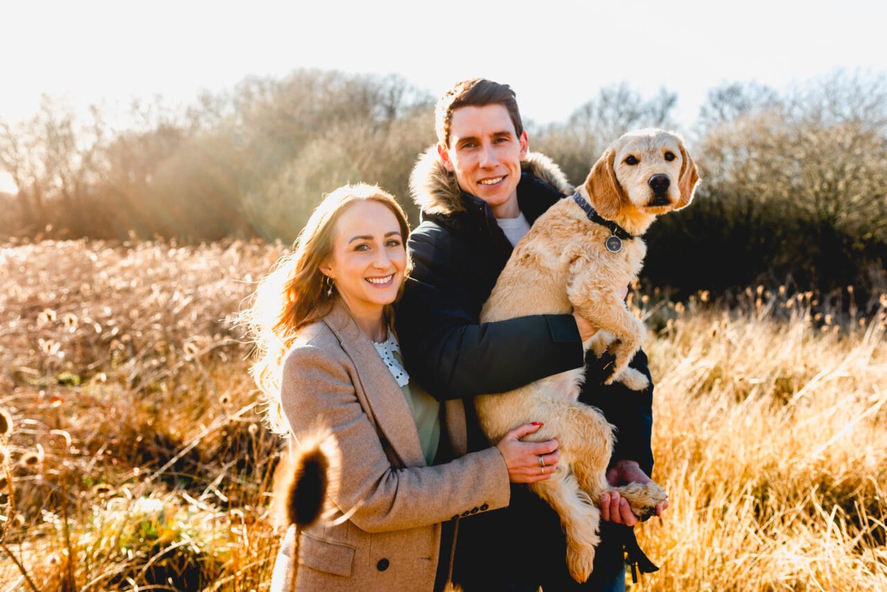 Engaged couple stand together in a field on long blonde grass with their dog, Otis in the mans arms.