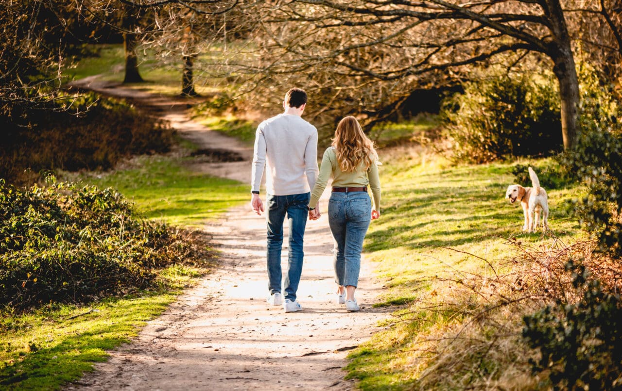 Man and woman walk away from the camera down a country path with a dog watching them from the pathway