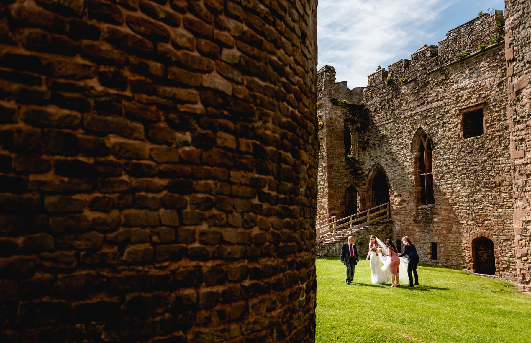 Bride and her Dad stand in the grounds of Ludlow Castle before the wedding ceremony. They are in the shade of the round centre building