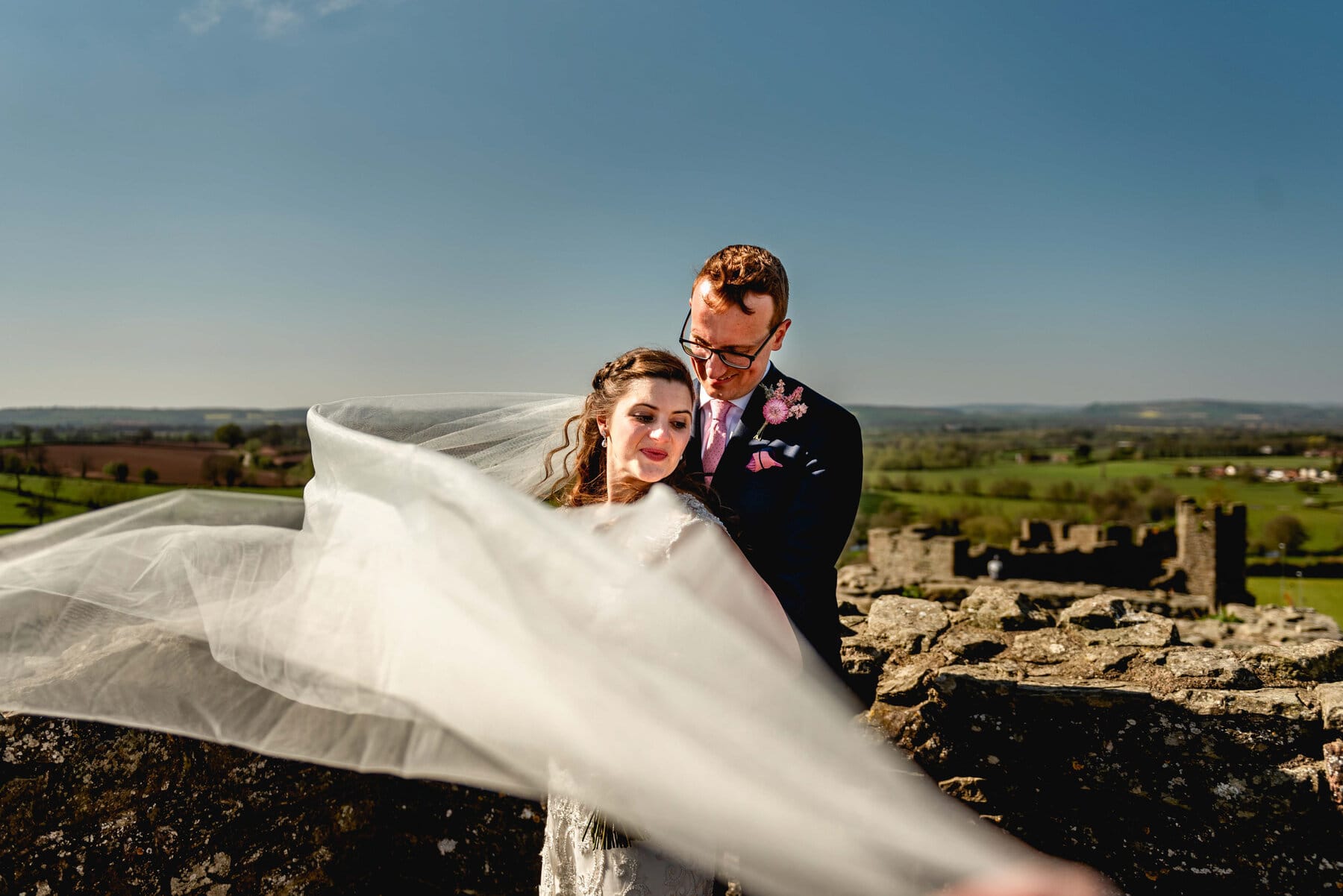 Bride and groom portrait, veil blows infant of bride as they stand on the top of a turret at Ludlow Castle with a blue sky behind the,, groom holds bride from behind.