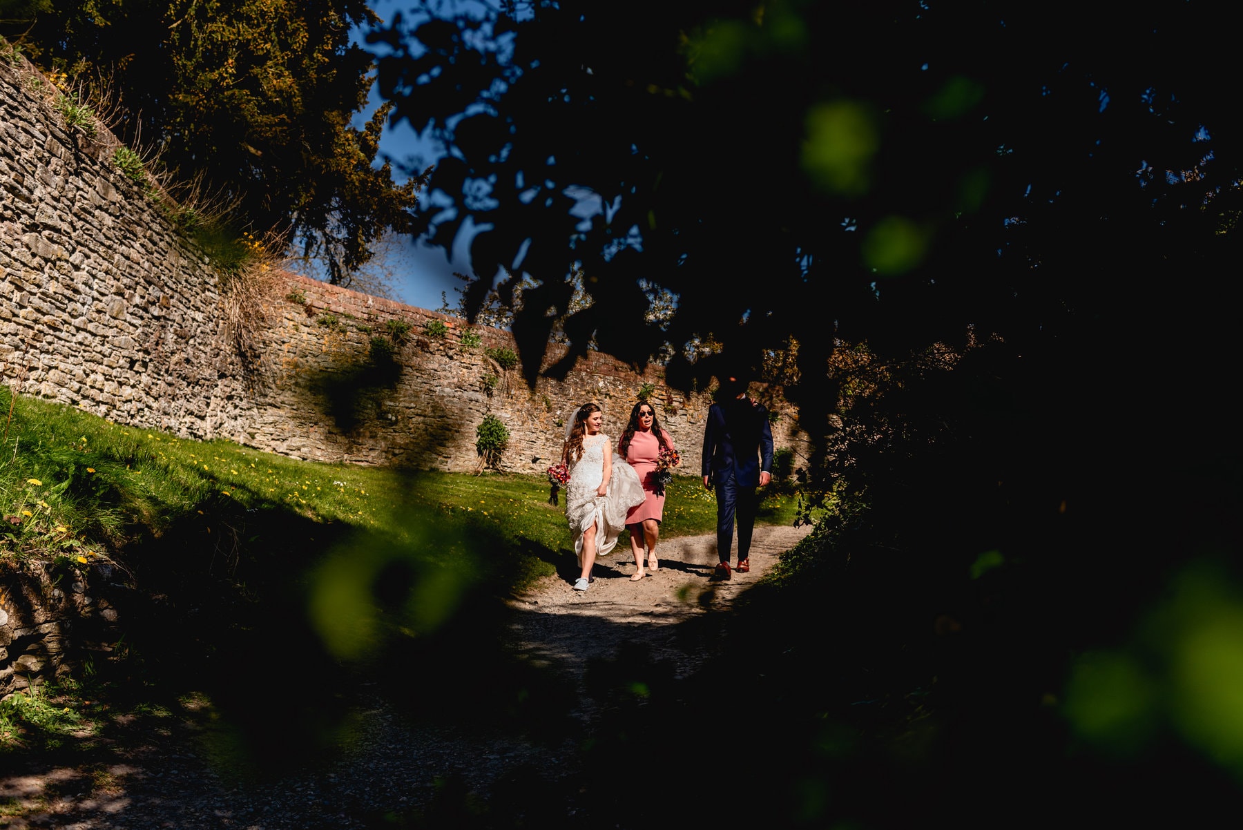 Bridal party are seen through the shadow of trees as they leave their wedding venue to walk the short distance to their wedding reception