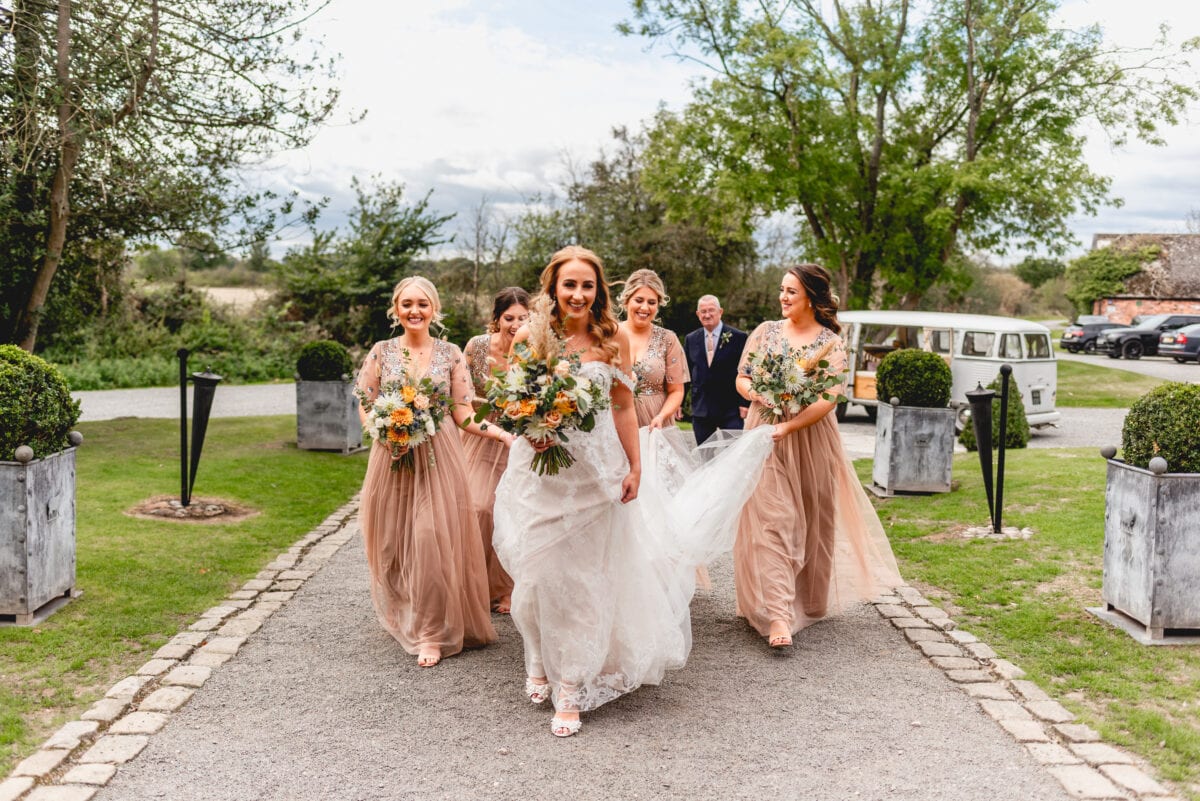 Bride and bridesmaids arrive at wedding venue, walk up path to Shustoke Barn holding dress