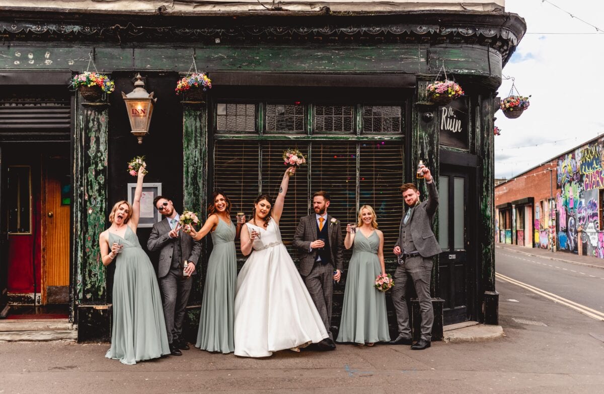 Bridal Party photograph with bride, bridesmaids and groomsmen all cheering with a drink in their hands standing on a street outside The Ruin pub in Digbeth